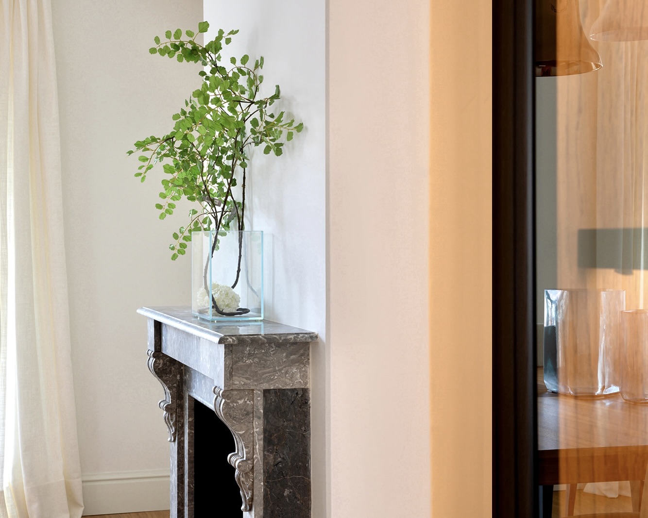 Chimenea de mármol con un jarrón de cristal conteniendo ramas verdes, creando una decoración elegante y natural en un interior luminoso con cortinas blancas y paredes claras.