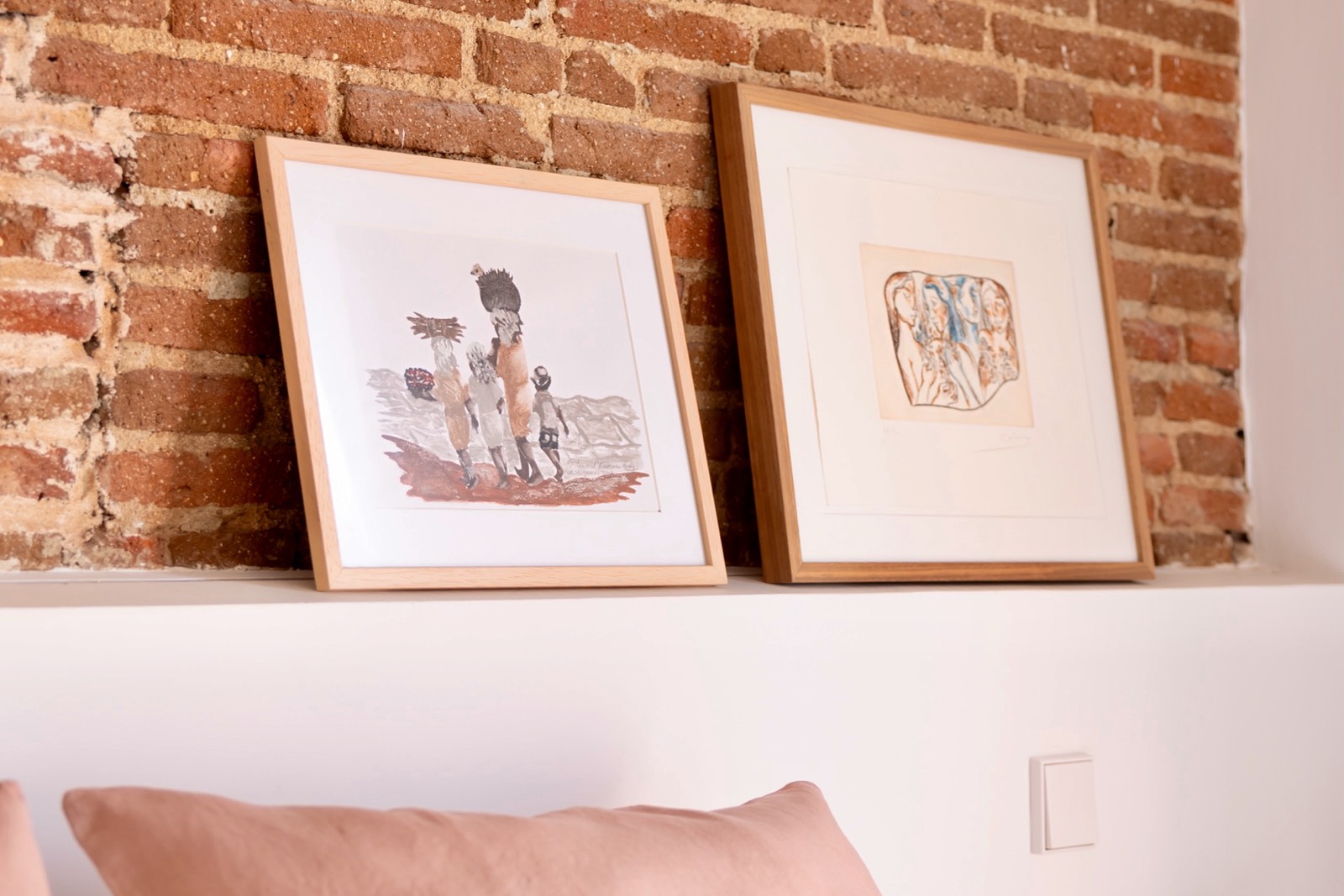 Dos marcos de madera con obras artísticas en tonos sepia y marrones, descansando sobre una repisa blanca frente a un muro de ladrillo rústico