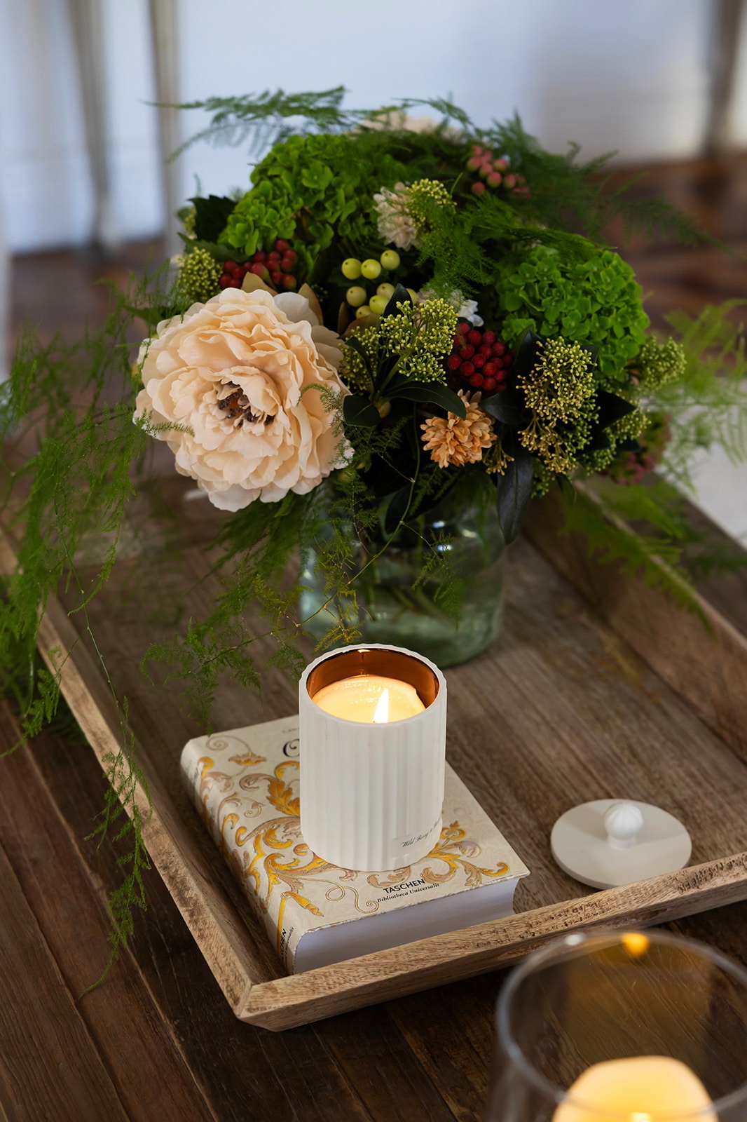 Arreglo floral con rosa durazno, flores verdes y bayas rojas en jarrón de cristal, junto a una vela aromática blanca encendida sobre un libro decorativo en una bandeja de madera rústica