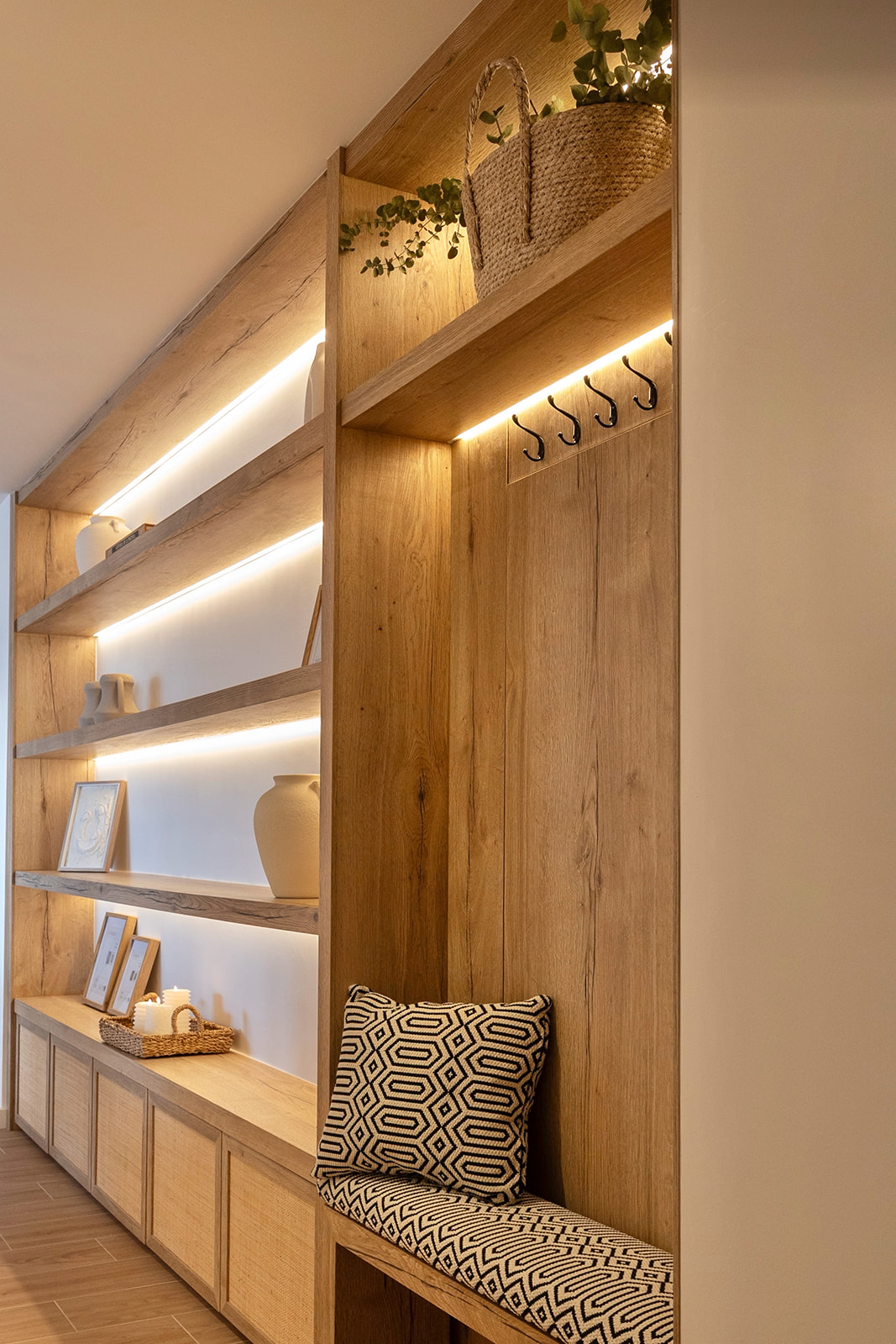 Recibidor moderno con mueble de madera empotrado, estanterías iluminadas con LED, percheros y un asiento acogedor con cojín estampado. Decorado con plantas y objetos decorativos para crear un ambiente cálido y acogedor.