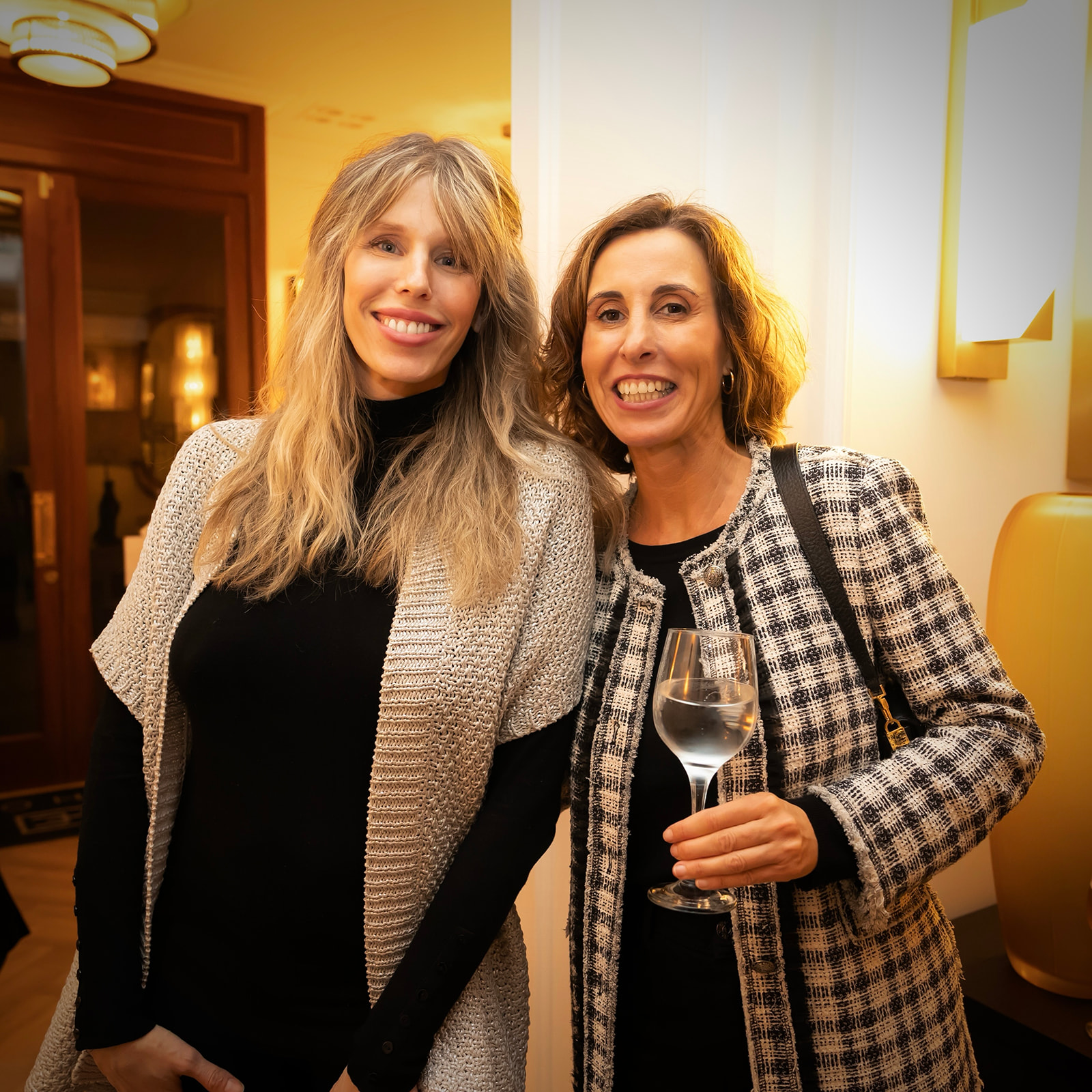 Dos mujeres sonrientes en un evento social. Una lleva un suéter gris sobre una prenda negra, la otra viste una chaqueta a cuadros y sostiene una copa de vino. El ambiente es cálido y acogedor.