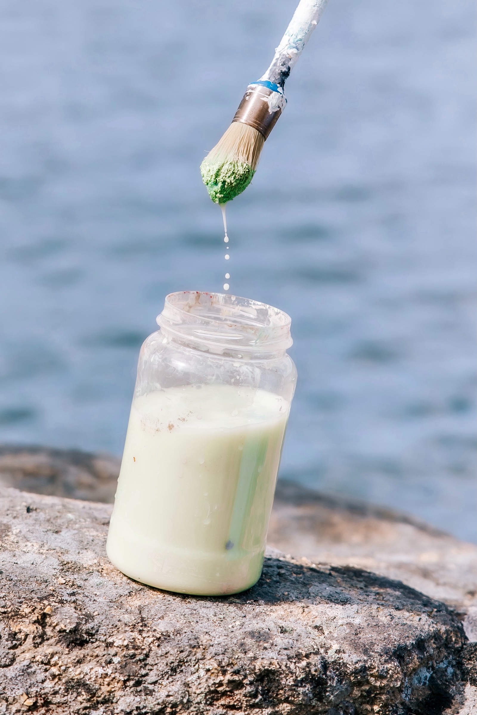 Un pincel con pintura verde goteando en un frasco de vidrio lleno de pintura blanca, colocado sobre una roca junto al mar azul