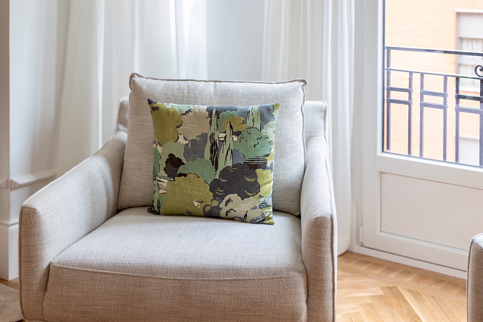 Sillón beige cómodo con un cojín decorativo de estampado verde y gris junto a una ventana con cortinas blancas y balcón con barandilla de hierro forjado. El suelo es de parquet y se ve parte de una pared naranja al fondo.