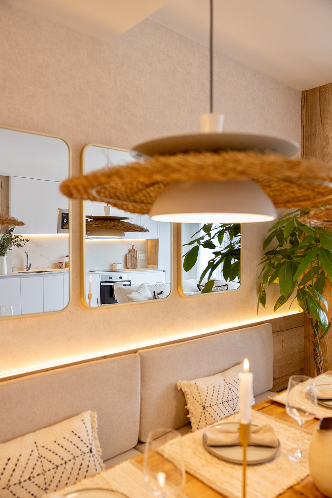 Comedor con lámpara de ratán y sofá beige, espejos redondos en la pared que reflejan una cocina blanca moderna. Ambiente cálido con tonos naturales y detalles bohemios.