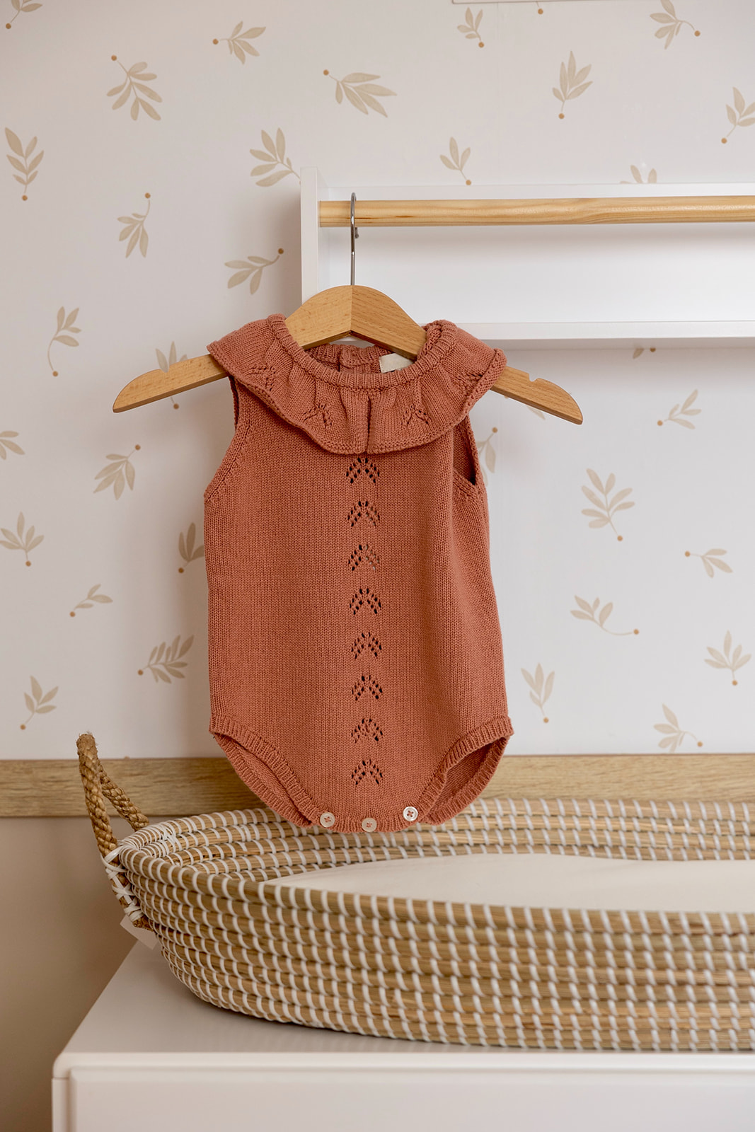 Body tejido de color terracota para bebé colgado en un perchero, con diseño de cuello volante y detalles calados. Está sobre un cambiador con un cesto de mimbre y papel tapiz floral de fondo.