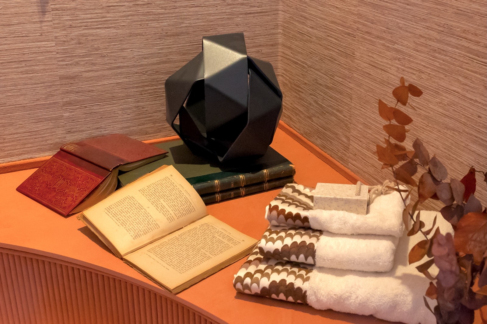 Composición de libros antiguos, toallas blancas con diseño, lámpara geométrica negra y rama seca sobre superficie naranja