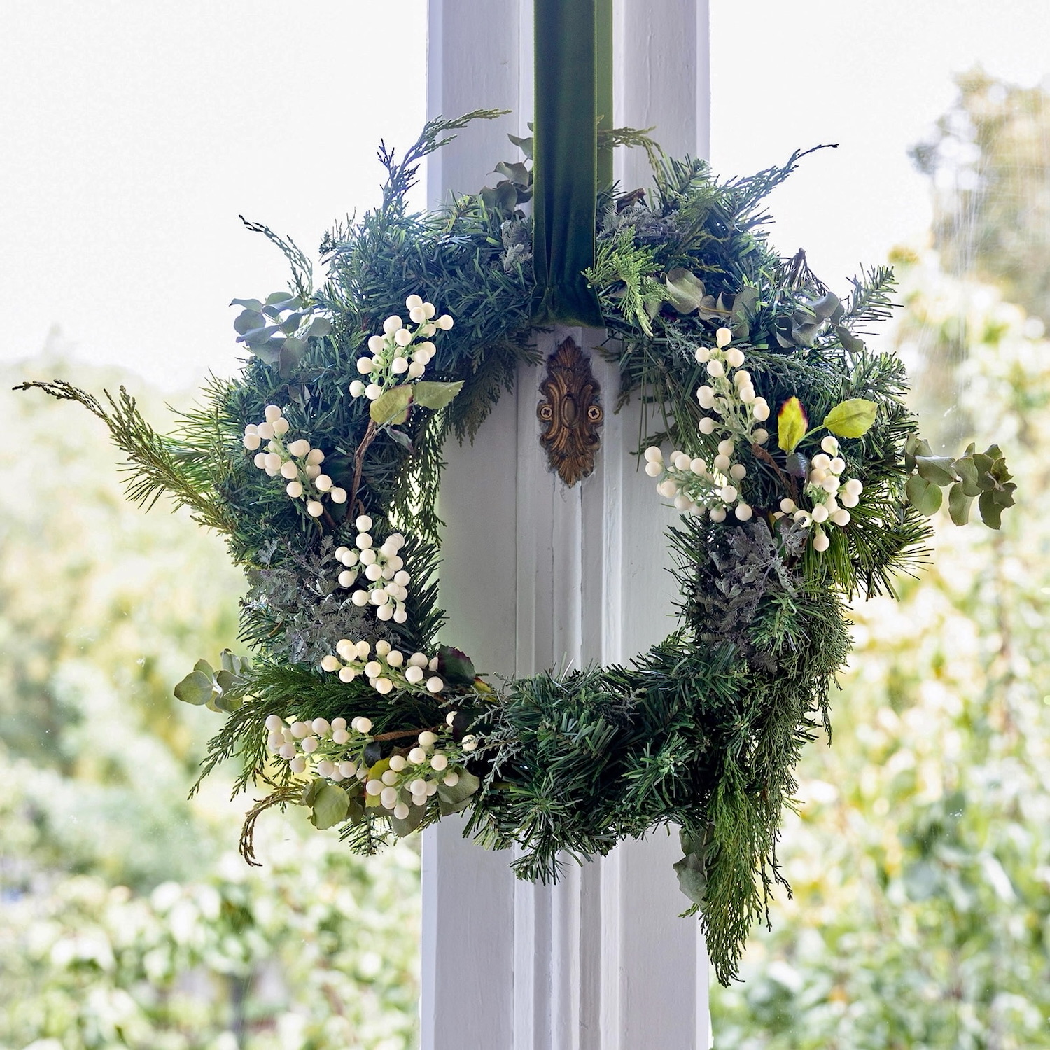Corona navideña colgada en una columna blanca, hecha con ramas de pino verde fresco y decorada con bayas blancas y hojas de eucalipto. La corona tiene un detalle ornamental de bronce en el centro de la columna.