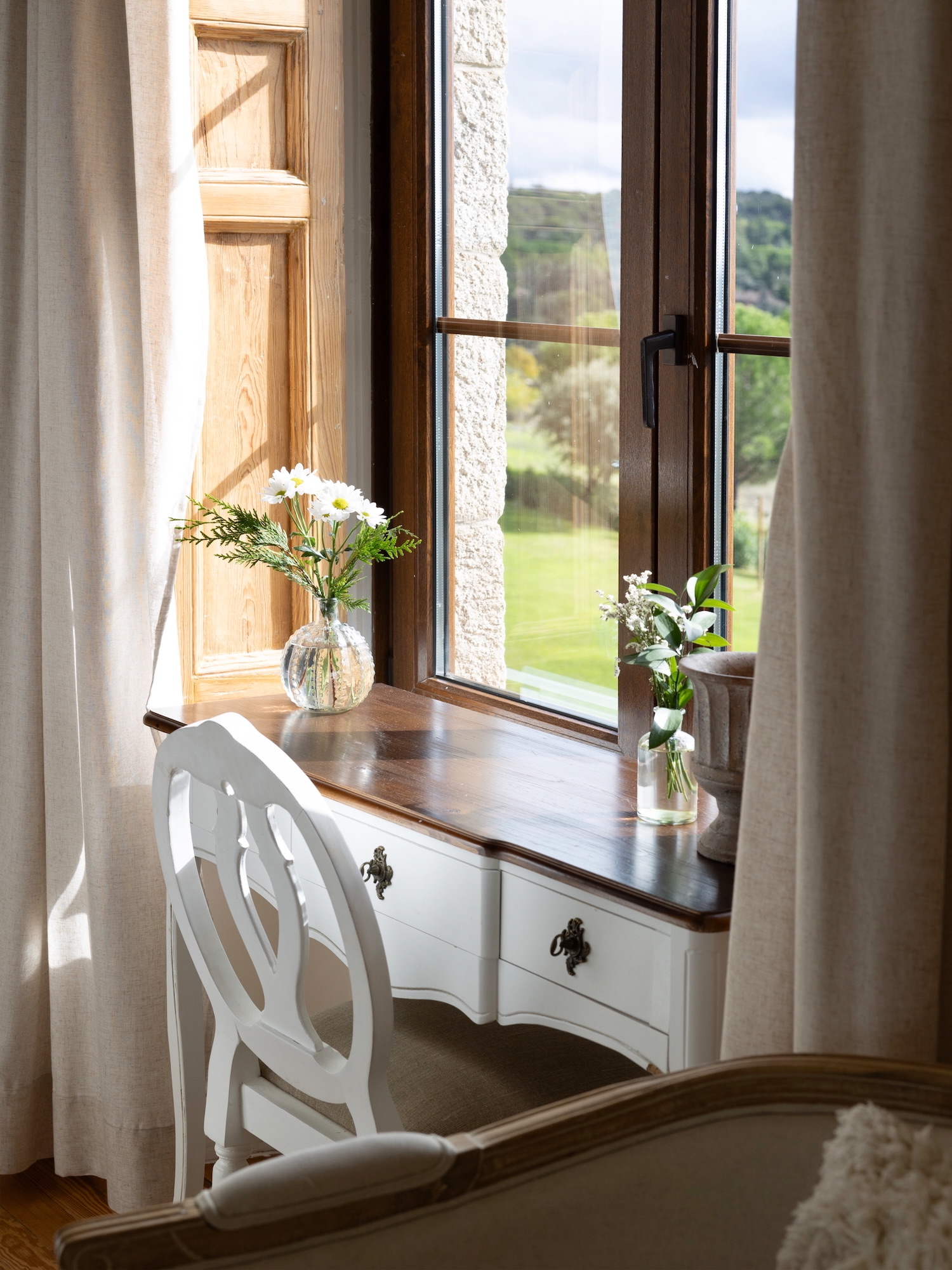 Escritorio vintage blanco con superficie de madera oscura junto a una ventana luminosa, decorado con floreros de cristal conteniendo margaritas blancas y otras flores. Las cortinas beige enmarcan la escena con vista al jardín.