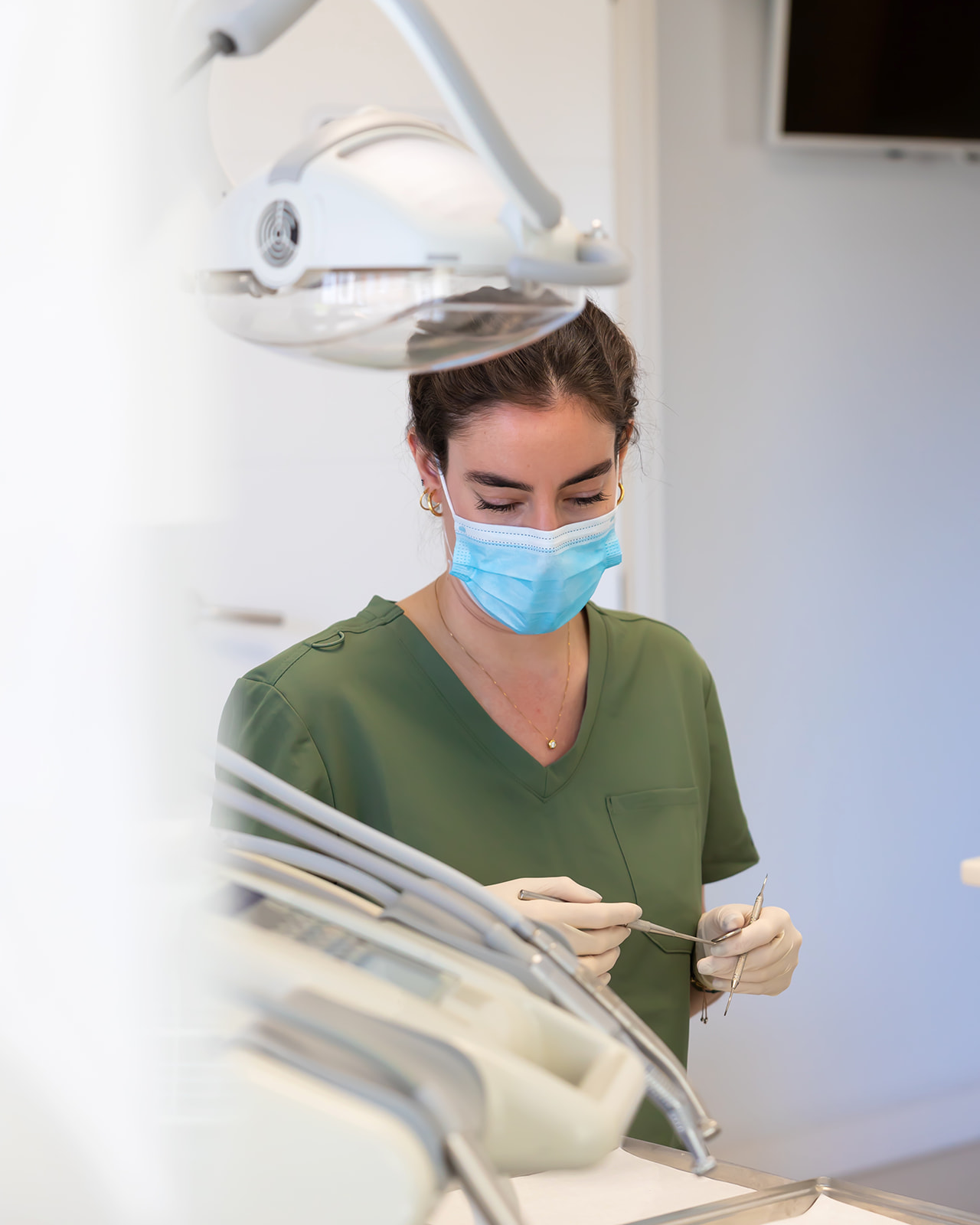 Profesional de la salud dental con uniforme verde y mascarilla azul trabajando en un consultorio odontológico moderno, con equipo dental visible en primer plano y fondo.