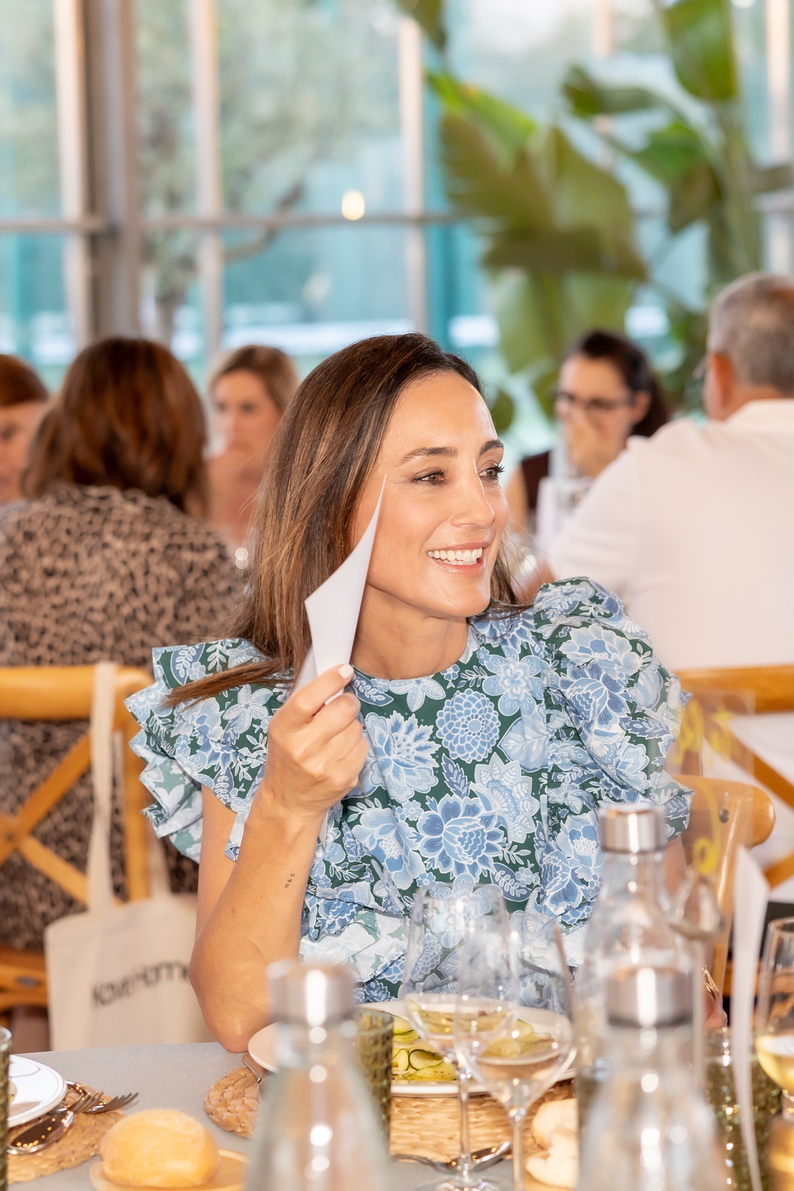 Mujer sonriente con blusa floral azul y blanca sentada en un evento social, sosteniendo una servilleta con mesa servida en primer plano