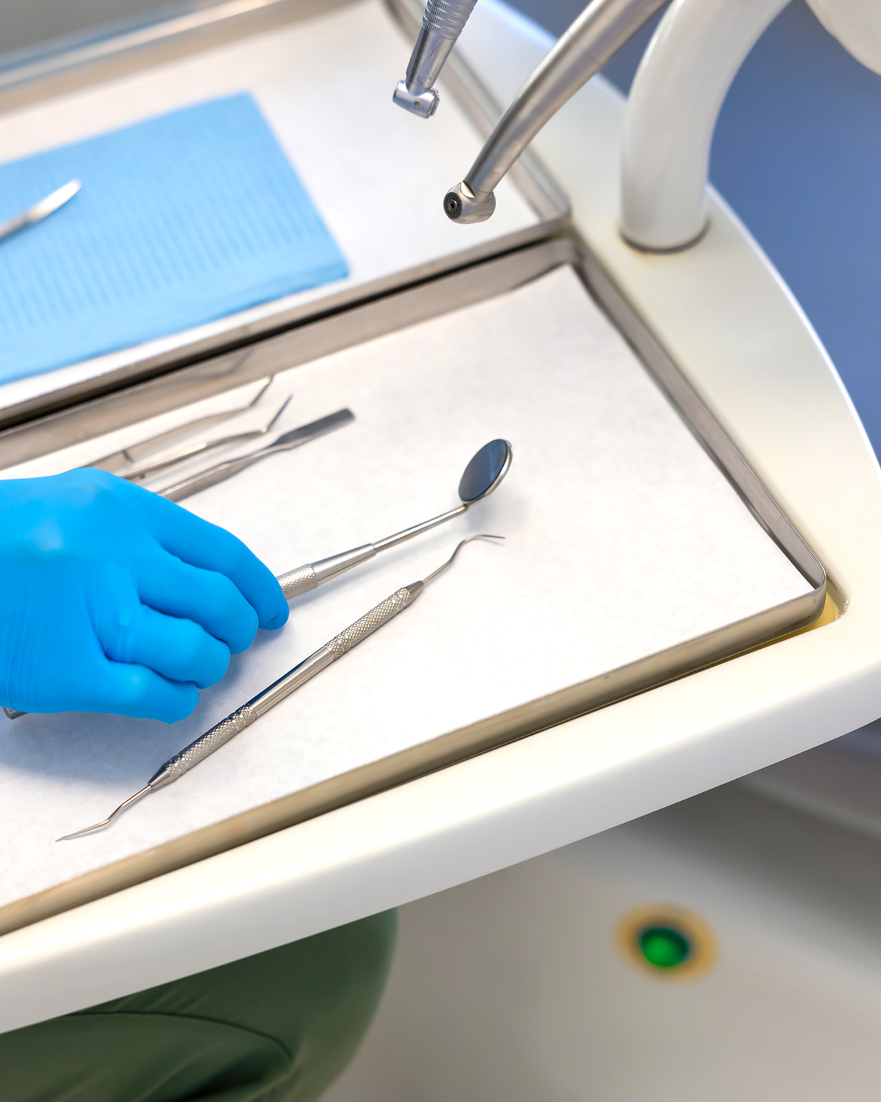 Imagen de instrumentos dentales en una bandeja metálica. Una mano con guante azul sostiene un espejo dental. Se ven otros instrumentos como pinzas y una pieza de mano dental.