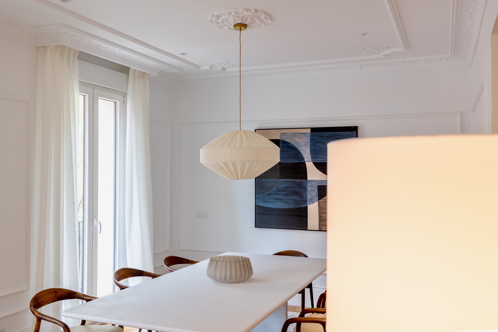 Comedor elegante con diseño minimalista. Mesa blanca rectangular con sillas de madera. Lámpara colgante moderna y cuadro abstracto azul en la pared. Techo con molduras decorativas y cortinas blancas en ventana.
