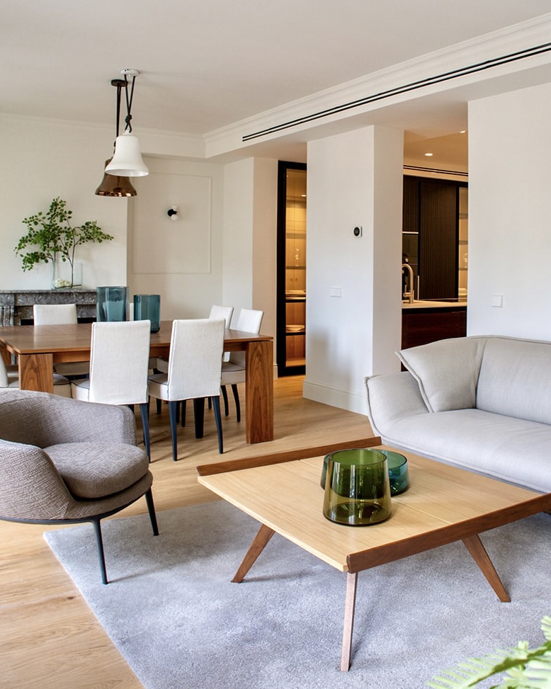 Sala de estar y comedor modernos con estilo minimalista. Muebles en tonos neutros, mesa de madera, sillas blancas, sofá gris claro y sillón. Detalles decorativos como plantas y vasos de colores sobre una mesa de centro de madera.