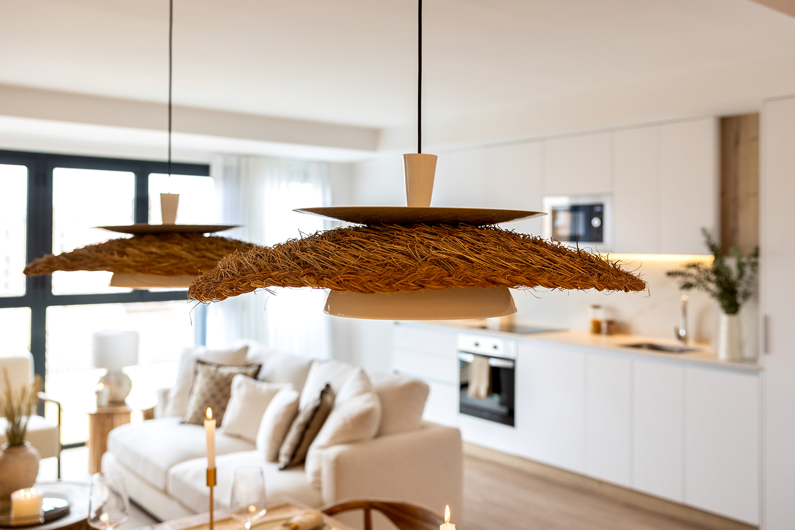 Salón y cocina abierta con lámparas colgantes de paja en primer plano. Decoración moderna y luminosa con sofá blanco y cocina integrada al fondo.