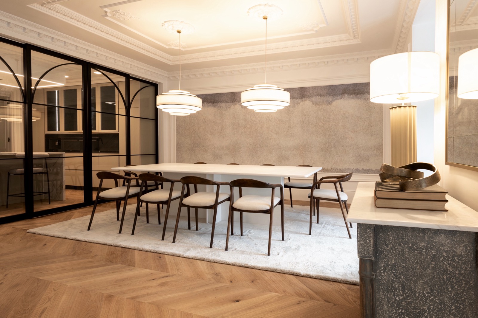 Elegante comedor moderno con mesa blanca larga, sillas de madera oscura, lámparas colgantes cilíndricas, techo ornamentado, pared de vidrio divisoria y piso de madera en espiga.
