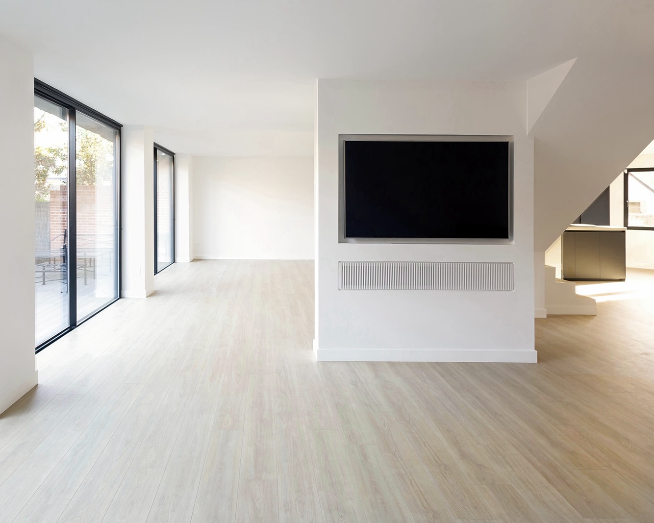 Interior de una sala moderna y minimalista con paredes blancas, piso de madera clara, ventanales grandes, y una televisión de pantalla plana montada en la pared. El espacio es luminoso y amplio.