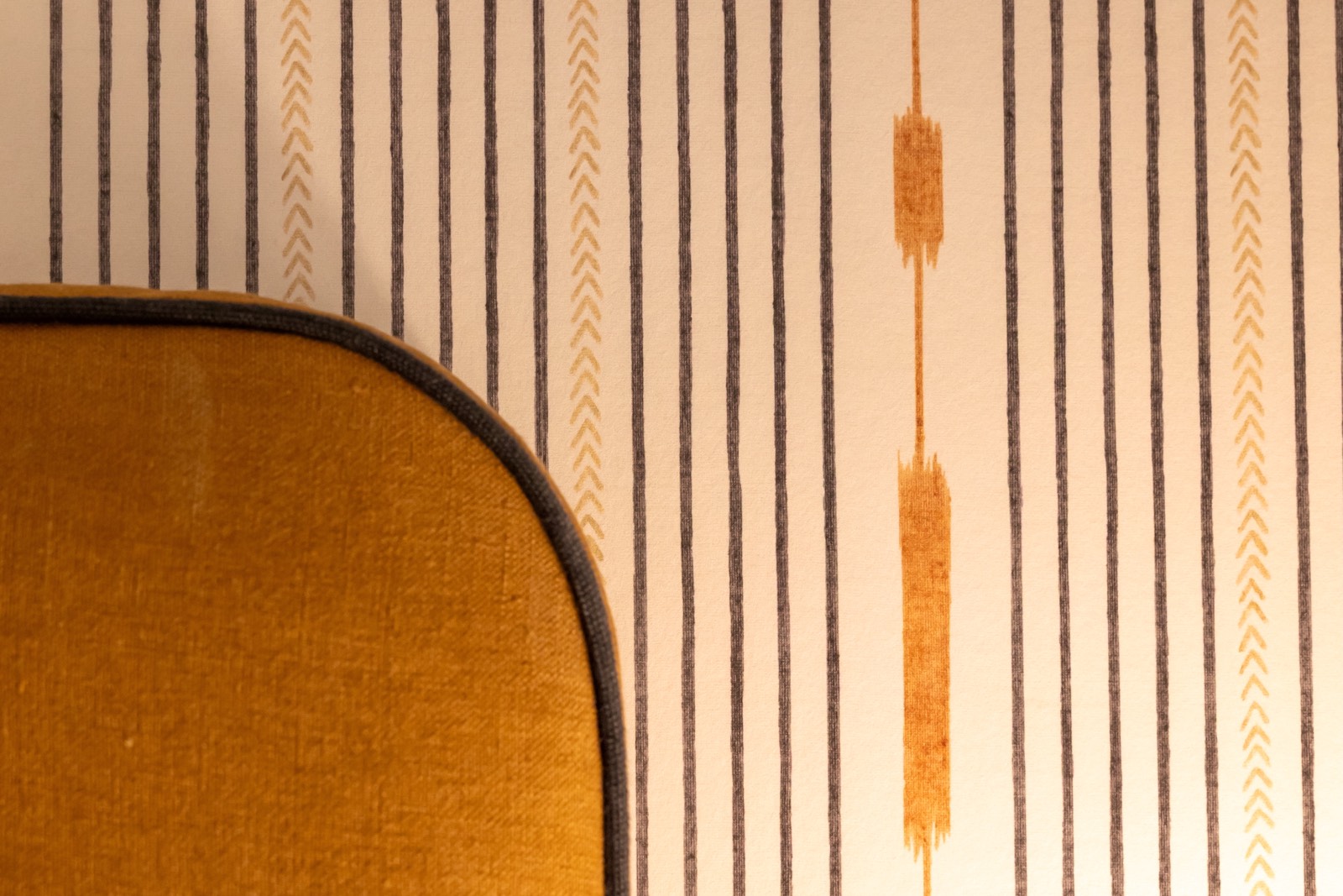 Detalle de una pared con papel tapiz a rayas verticales en tonos beige y gris, con dos elementos naranjas verticales. En primer plano se ve una esquina de un sillón de color mostaza.