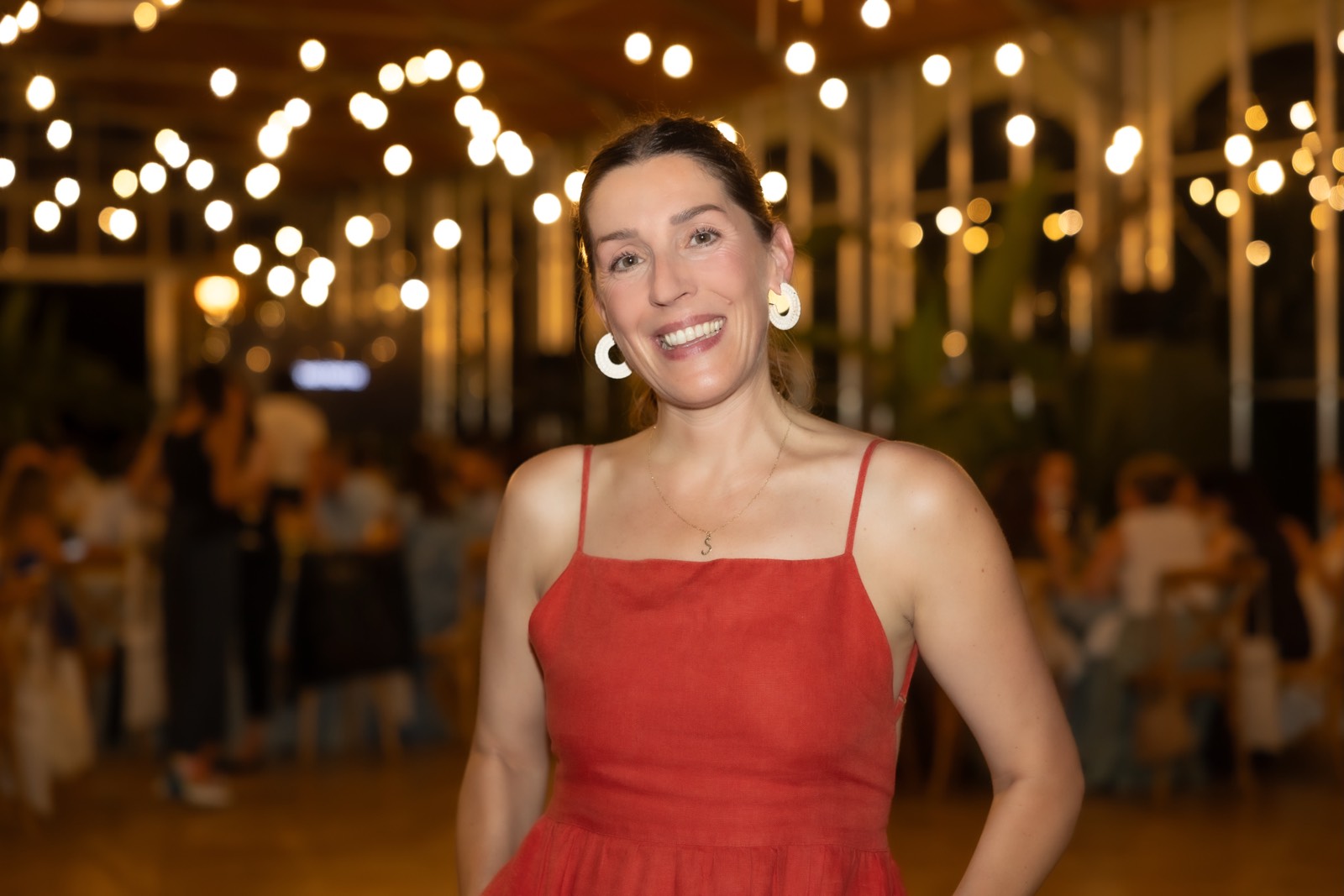 Mujer sonriente con vestido rojo de tirantes, usando aretes blancos, en un ambiente festivo con luces decorativas desenfocadas