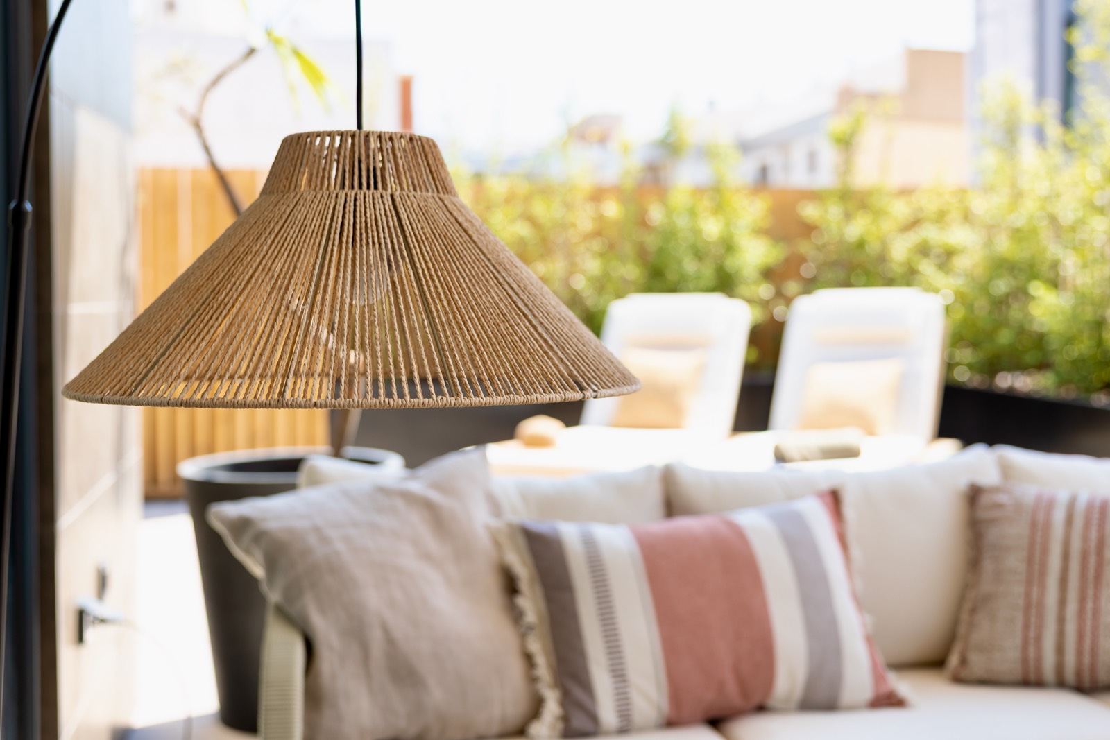 Terraza acogedora con una lámpara de mimbre colgante sobre un sofá con cojines coloridos. Al fondo se ven sillas blancas y plantas verdes en un balcón soleado.