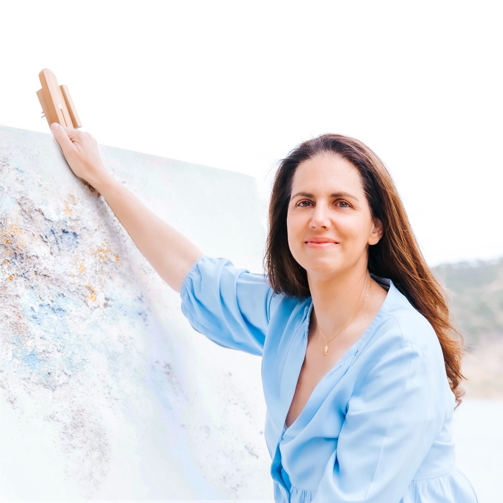 Mujer sonriente con blusa azul pintando un gran lienzo blanco al aire libre. Sostiene un pincel en la mano mientras trabaja en el lienzo apoyado contra una superficie. El fondo sugiere un entorno natural.