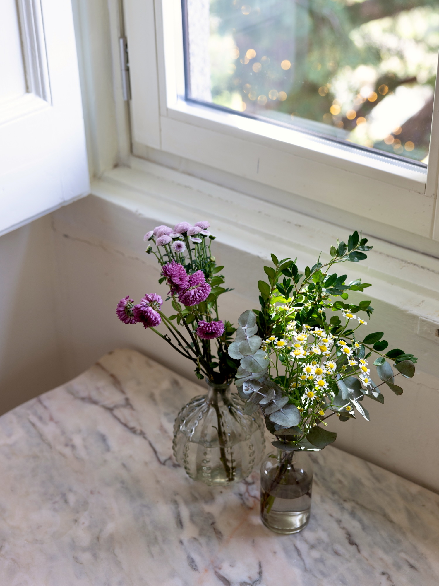 Dos jarrones de cristal con arreglos florales naturales sobre superficie de mármol junto a una ventana. Un jarrón contiene crisantemos morados y el otro margaritas blancas con eucalipto.