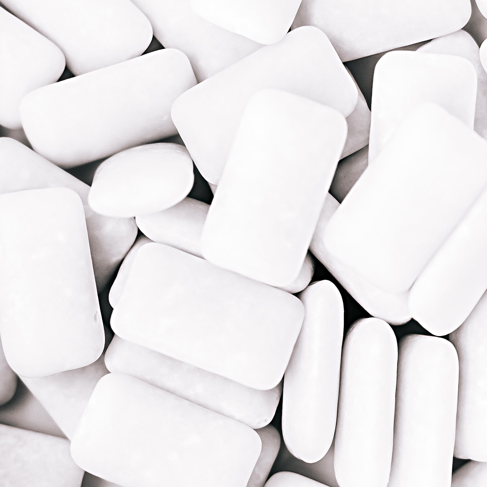 Primer plano de una colección de chicles blancos de diferentes formas y tamaños. Los chicles están amontonados y llenan todo el encuadre, creando un patrón abstracto en blanco y negro.