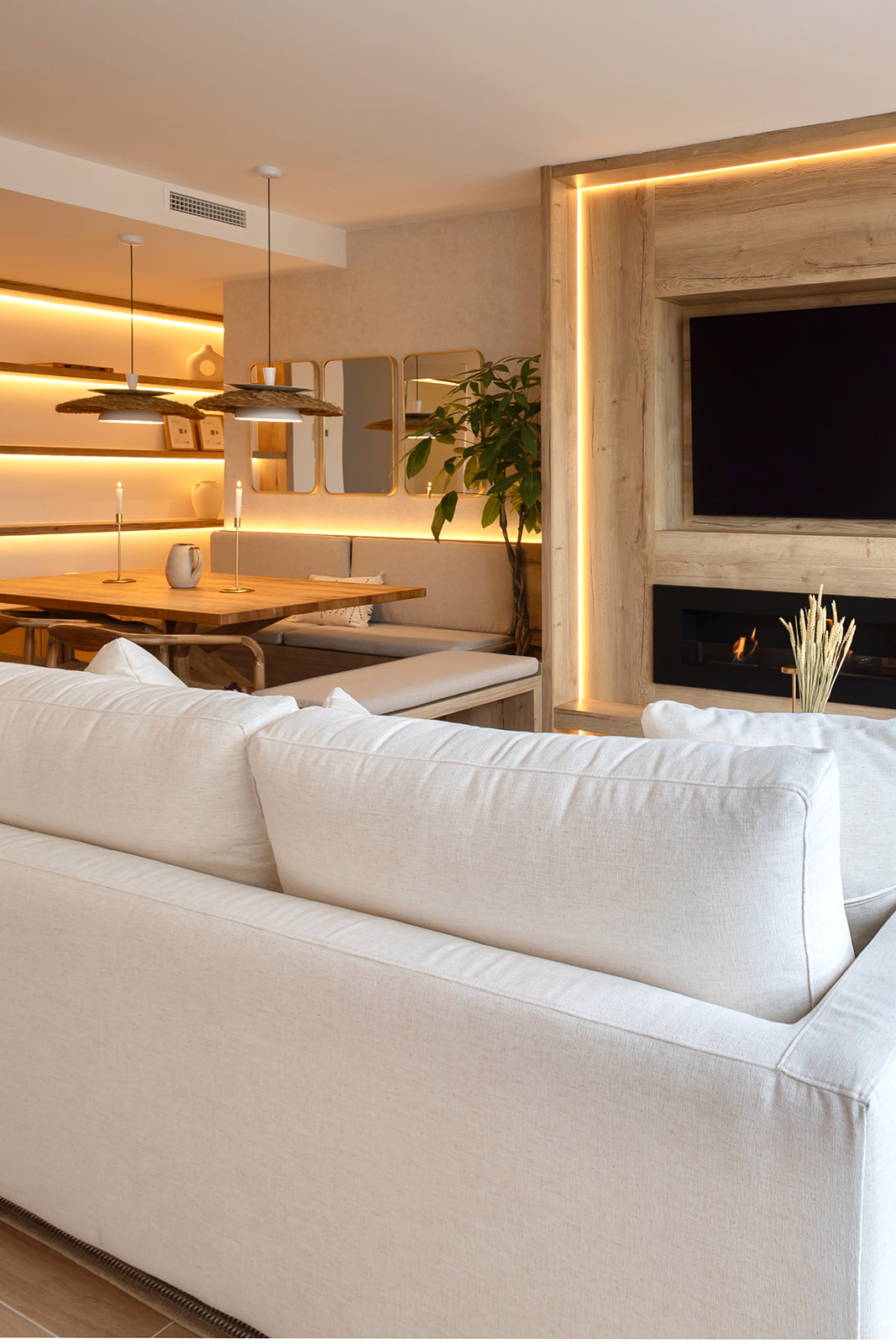 Sala de estar moderna con iluminación ambiental cálida, sofá blanco, comedor de madera, plantas decorativas y chimenea con televisor integrado en marco de madera