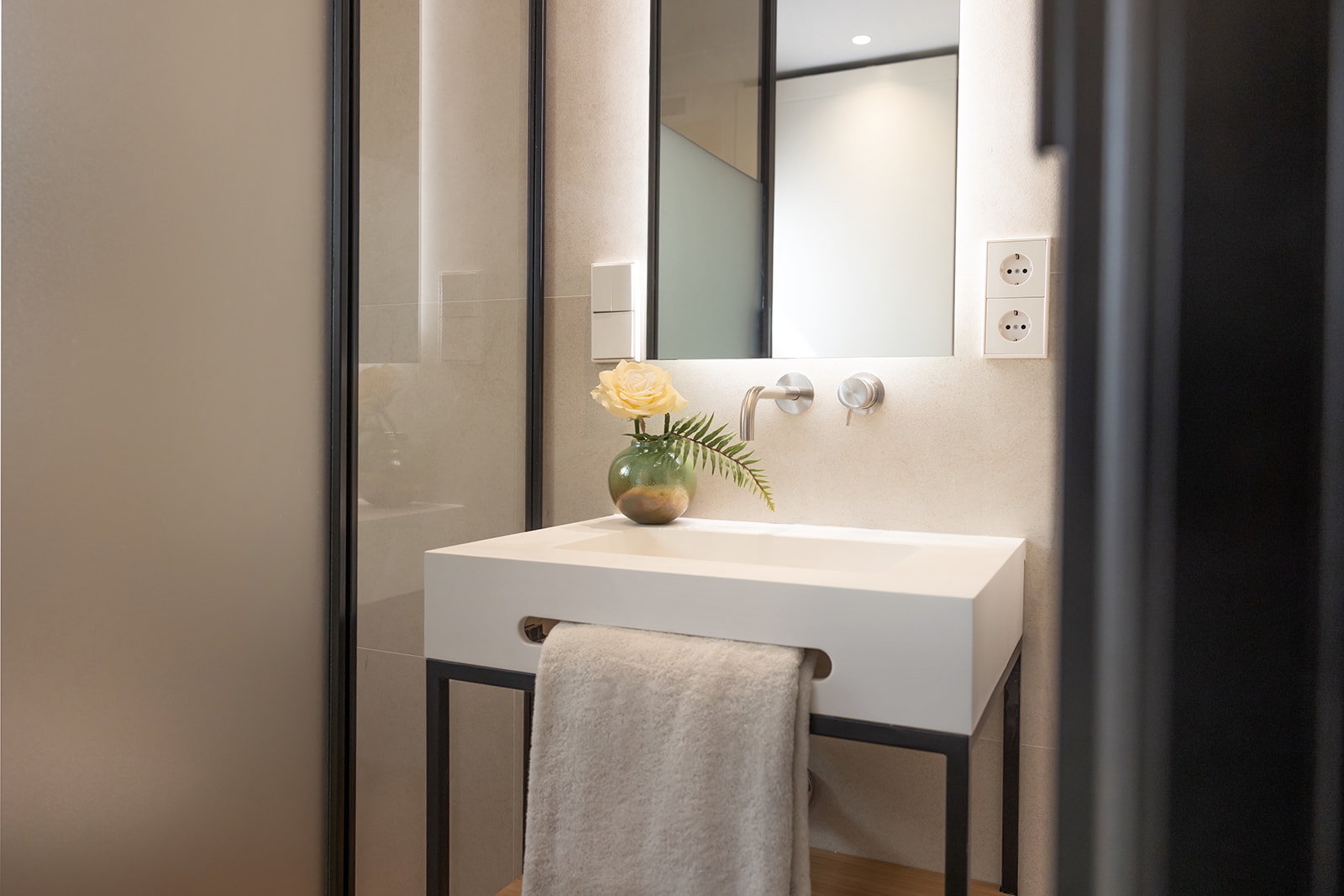Baño moderno y elegante con lavabo blanco sobre estructura metálica negra, espejo grande iluminado, flores en un jarrón y toalla blanca colgada. Detalles en tonos neutros y acabados limpios.