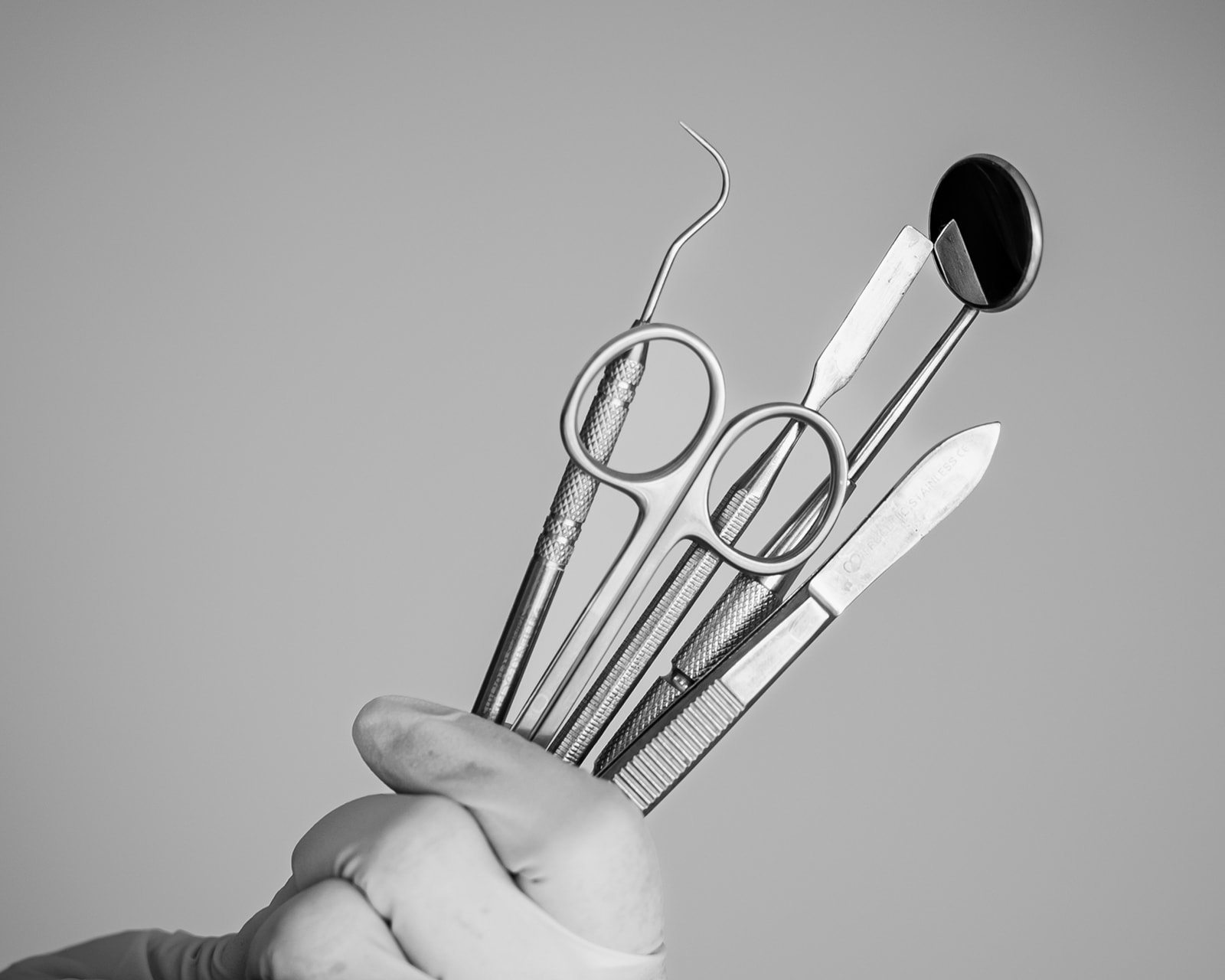 Imagen en blanco y negro de una mano sosteniendo varios instrumentos dentales, incluyendo un espejo, tijeras, un explorador y otras herramientas dentales contra un fondo gris claro.