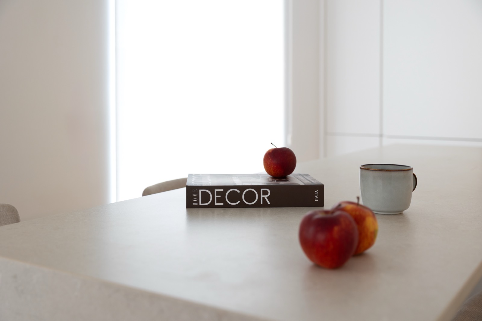 Mesa blanca con un libro de decoración, tres manzanas rojas y una taza de café blanca. La escena está iluminada por luz natural proveniente de una ventana, creando un ambiente minimalista y tranquilo.