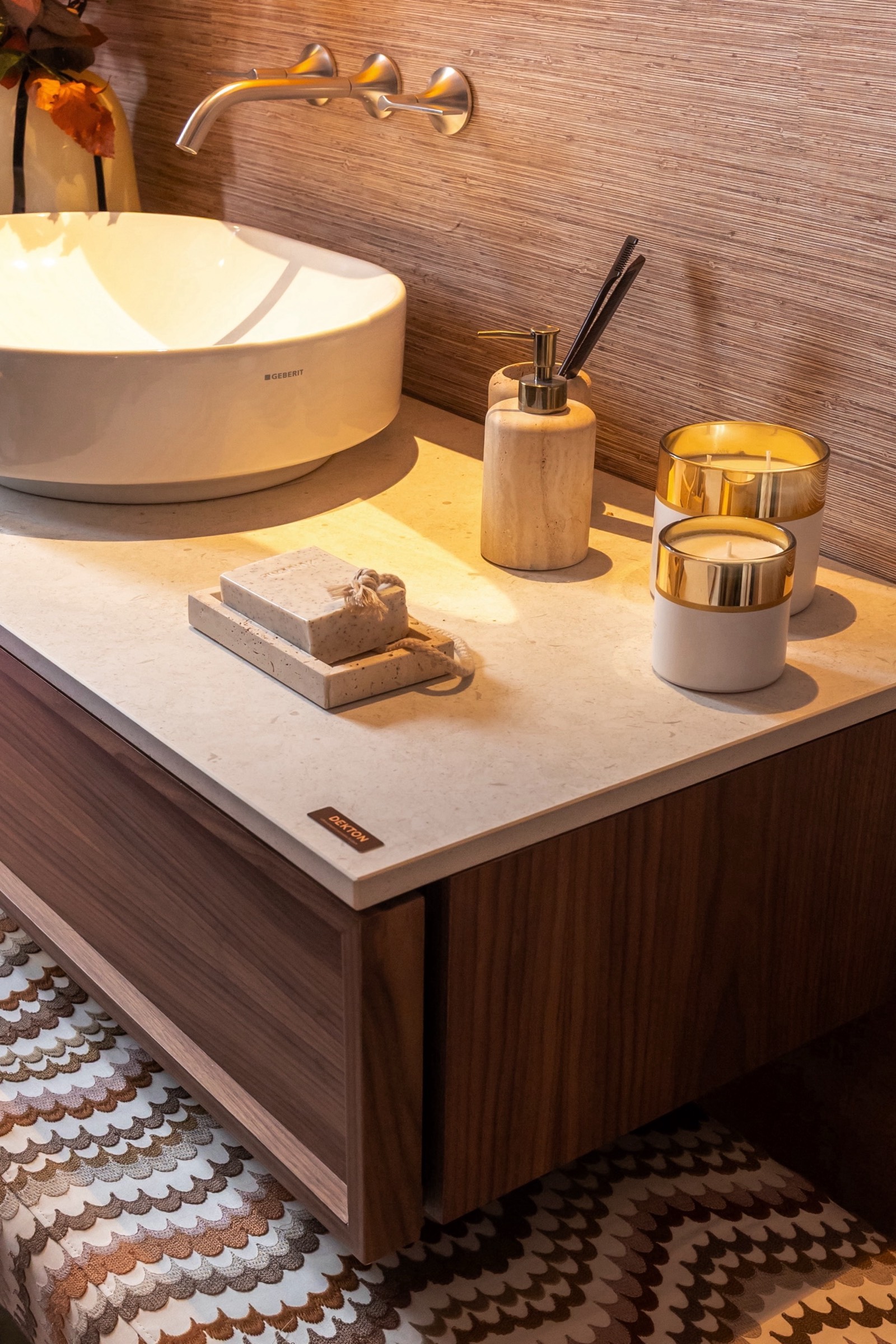 Detalle de lavabo moderno con lavamanos blanco circular, velas doradas, dispensador de jabón de madera y grifo dorado