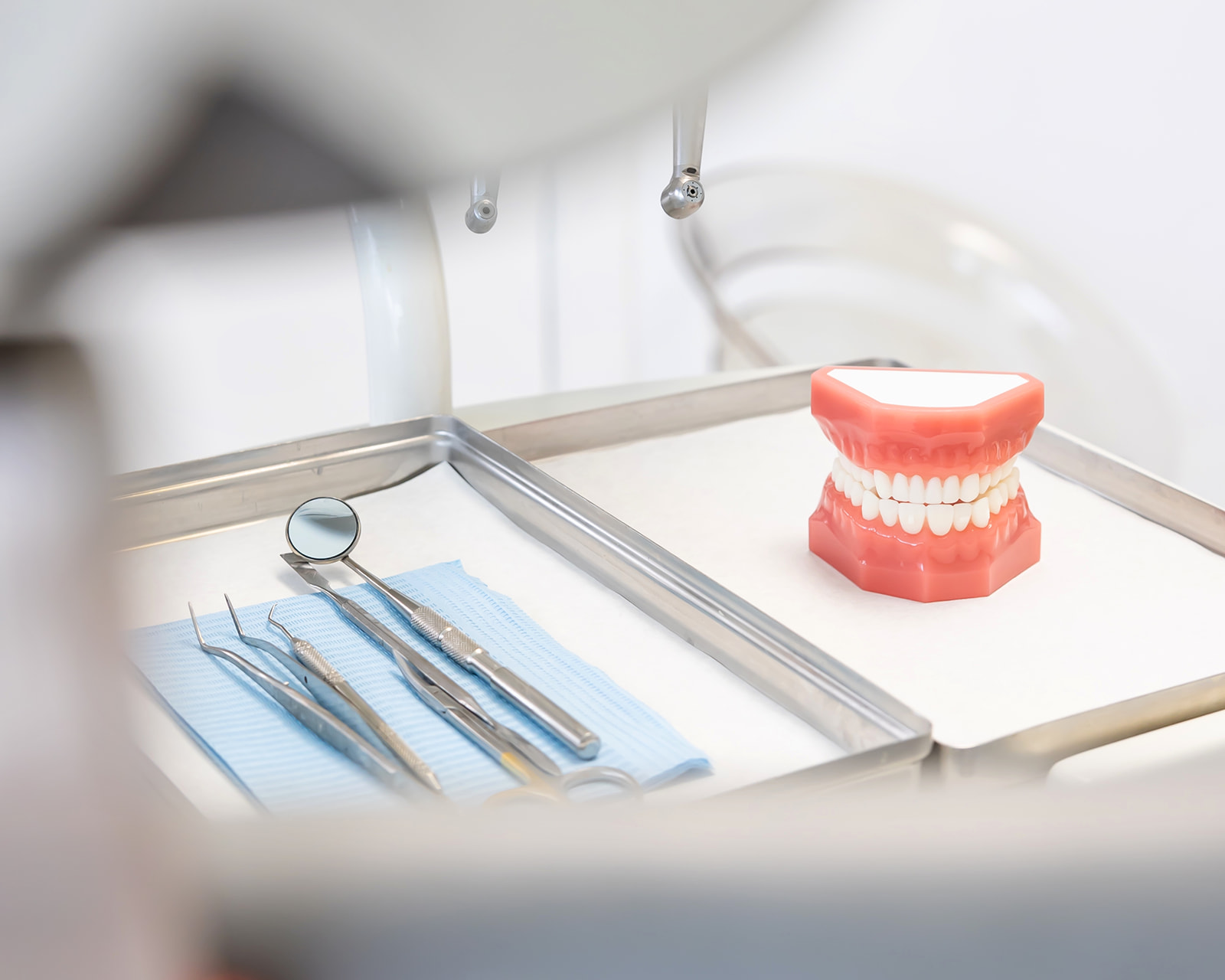 Bandeja de instrumentos dentales con espejo, pinzas y sonda junto a un modelo de dentadura en una clínica odontológica. Los instrumentos están sobre una tela azul y el modelo de dientes es de color rosa con dientes blancos.