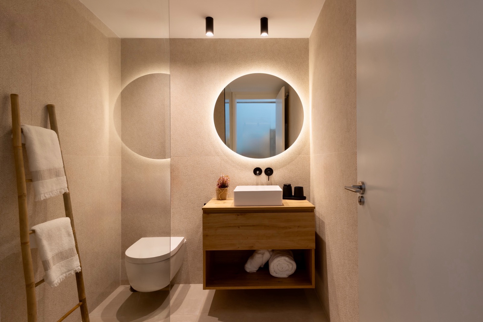 Baño moderno y minimalista con espejo redondo iluminado, mueble de madera con lavabo blanco, inodoro suspendido y escalera decorativa con toallas. Iluminación cálida y ambiente relajante.