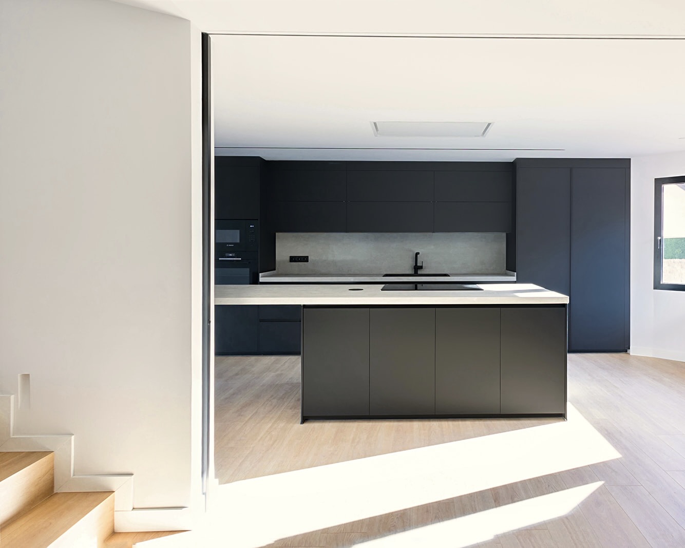 Cocina moderna y minimalista con muebles gris oscuro, isla central, encimera blanca y electrodomésticos integrados. Suelo de madera clara y paredes blancas que contrastan con el diseño oscuro de la cocina.