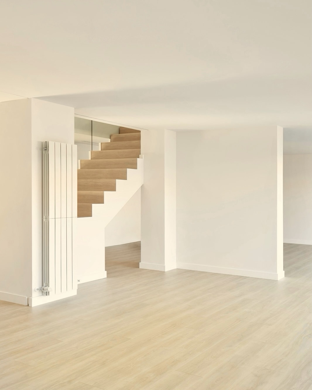 Interior minimalista con paredes blancas y suelo de madera clara. Una escalera de madera con diseño moderno se integra en la pared. Un radiador blanco vertical se encuentra junto a la escalera.