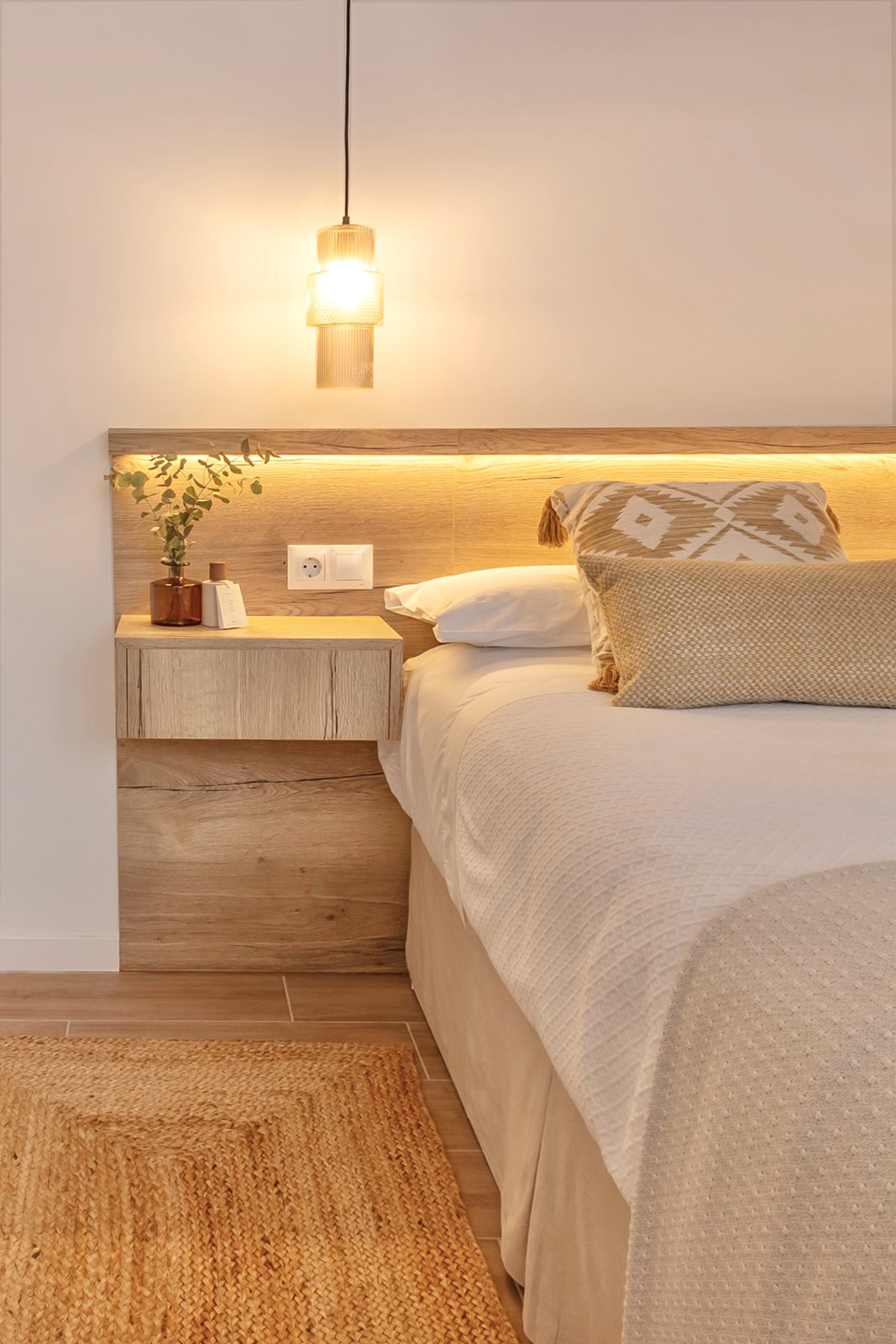 Dormitorio acogedor con iluminación cálida, cabecera de madera con luz integrada, mesita de noche incorporada, cama con ropa de cama blanca y cojines decorativos, lámpara colgante y alfombra de yute