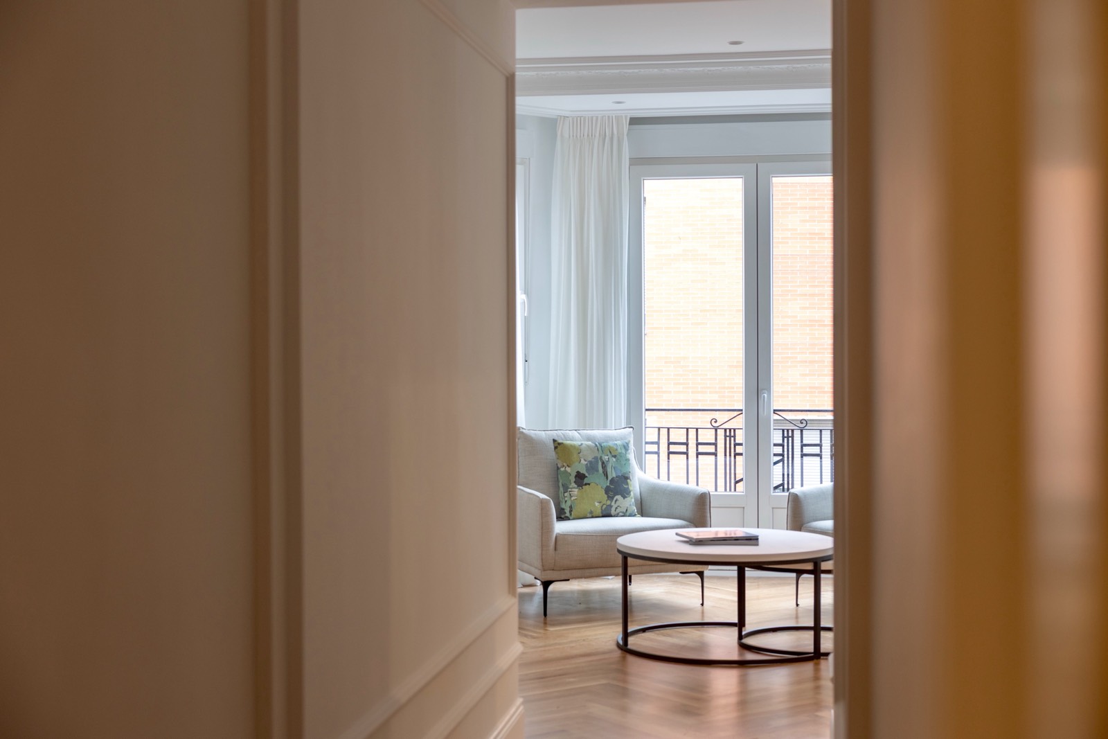 Vista de una sala de estar luminosa a través de una puerta abierta. La habitación tiene sillones claros, una mesa de centro redonda y un gran ventanal con cortinas blancas que da a un balcón.