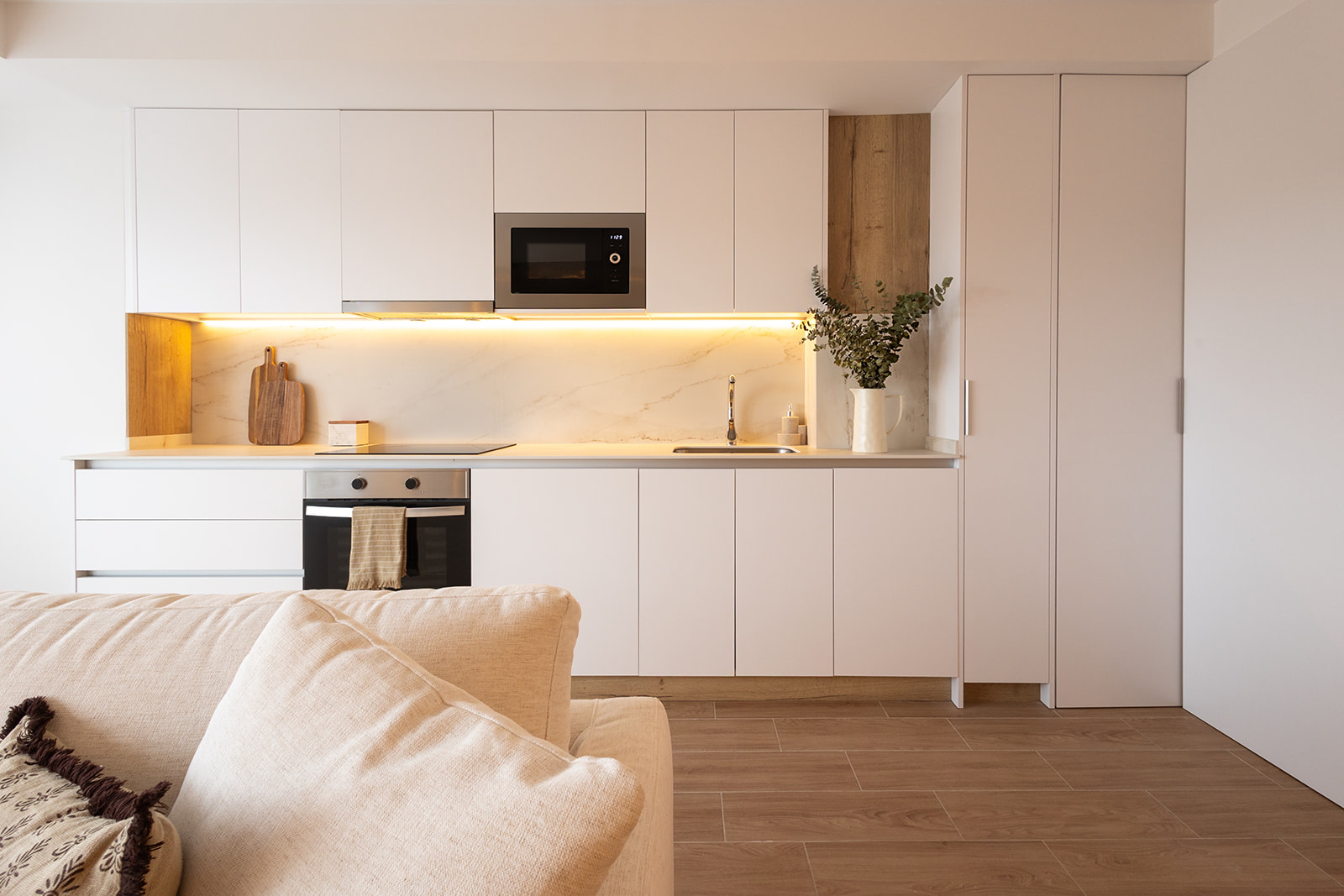 Cocina moderna y minimalista con gabinetes blancos, encimera clara, iluminación bajo los armarios superiores, electrodomésticos integrados y un sofá beige en primer plano. La decoración incluye una tabla de cortar y un jarrón con plantas.