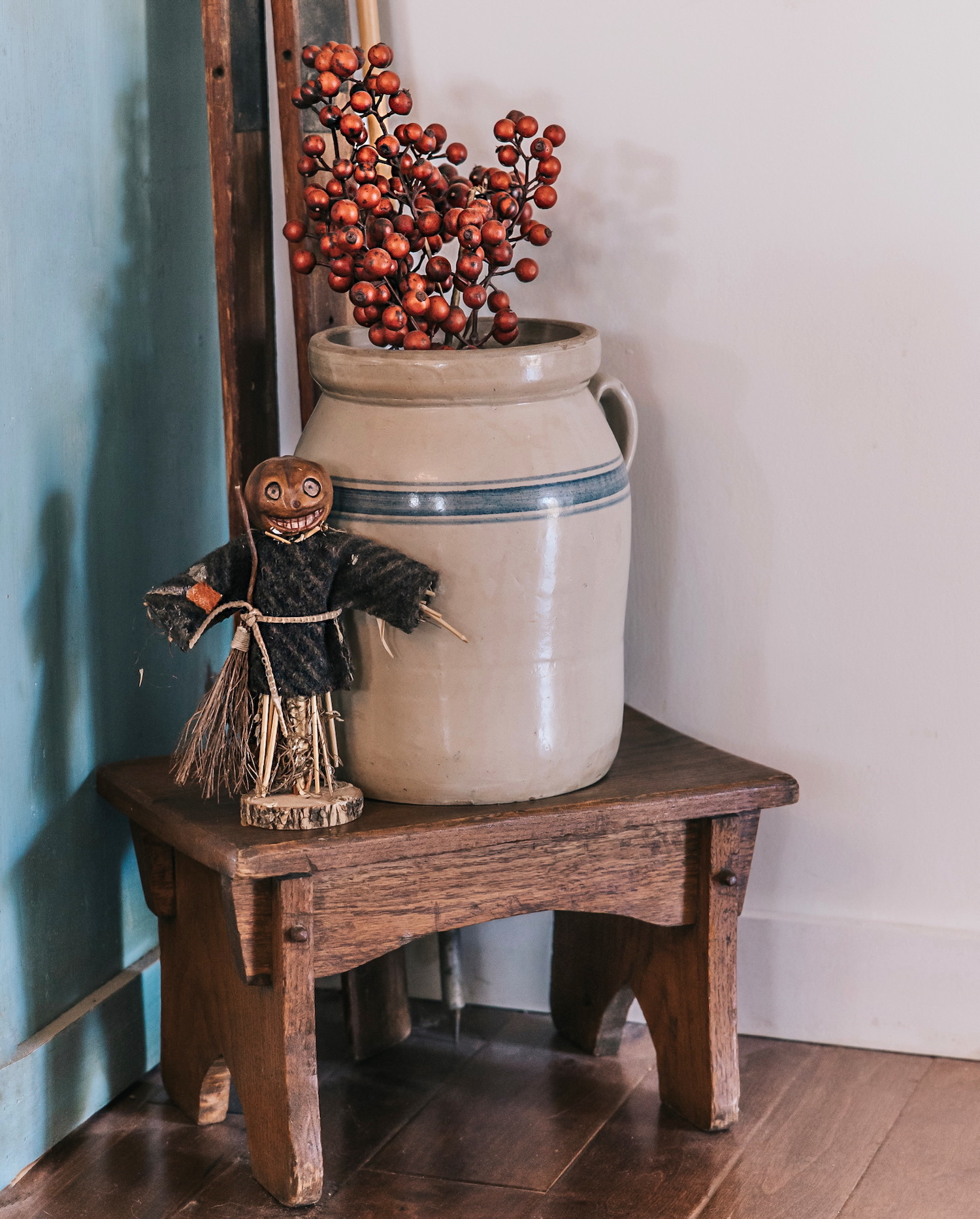 Decoración otoñal vintage con jarrón de cerámica beige conteniendo bayas rojas artificiales, junto a un pequeño espantapájaros decorativo sobre un banco rústico de madera