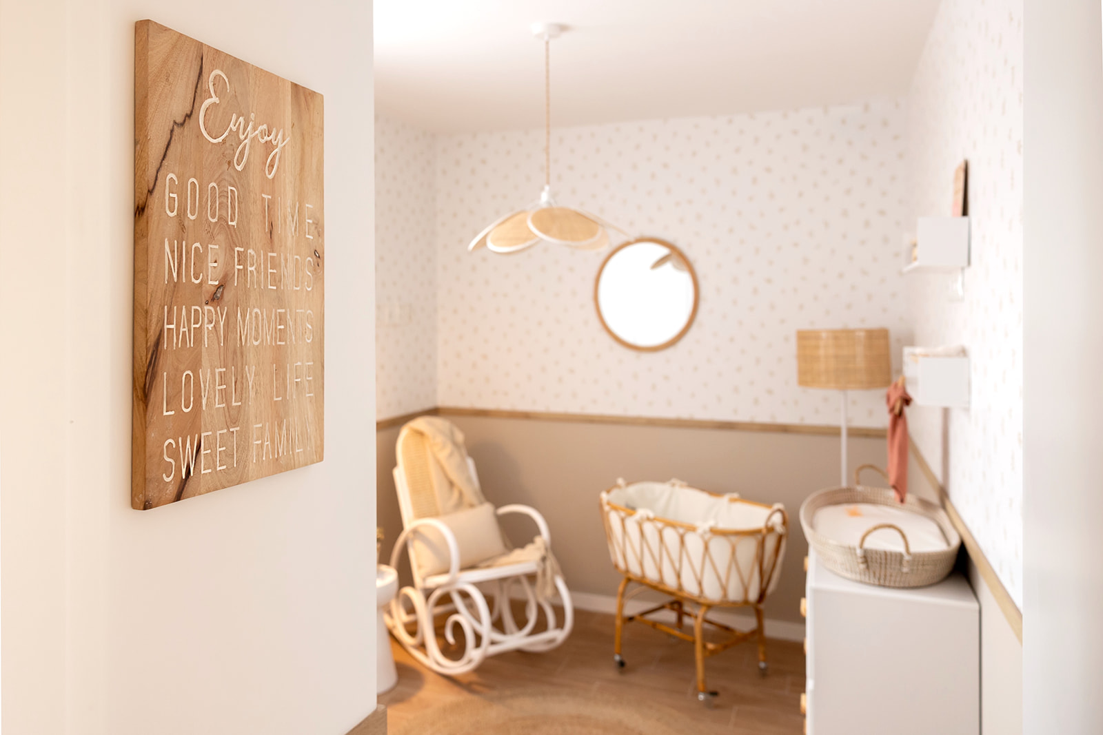 Habitación de bebé con estilo boho, decorada en tonos cálidos. Incluye una cuna de mimbre, una mecedora blanca, una lámpara colgante en forma de flor y un cartel de madera con un mensaje inspirador.