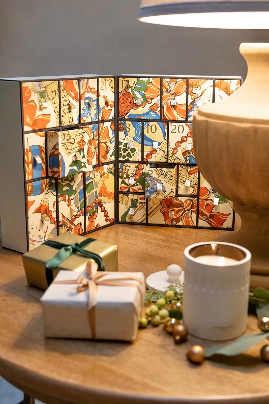 Calendario de adviento con diseño colorido y artístico junto a regalos envueltos, una vela blanca y decoraciones navideñas sobre una mesa de madera bajo una lámpara