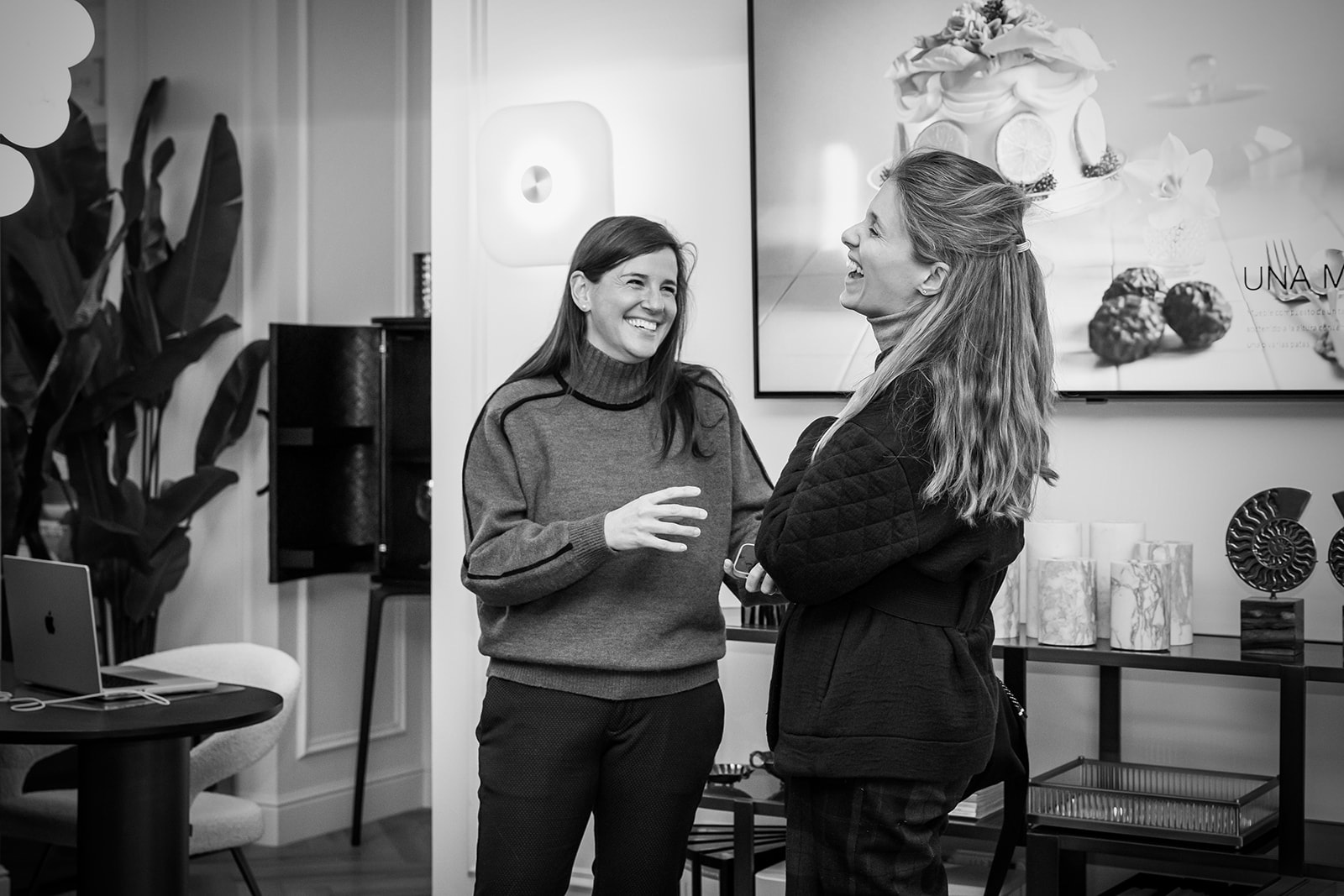 Imagen en blanco y negro de dos mujeres sonriendo y conversando en una oficina moderna. La habitación está decorada con plantas, cuadros y objetos decorativos. Hay un ambiente cálido y amistoso.
