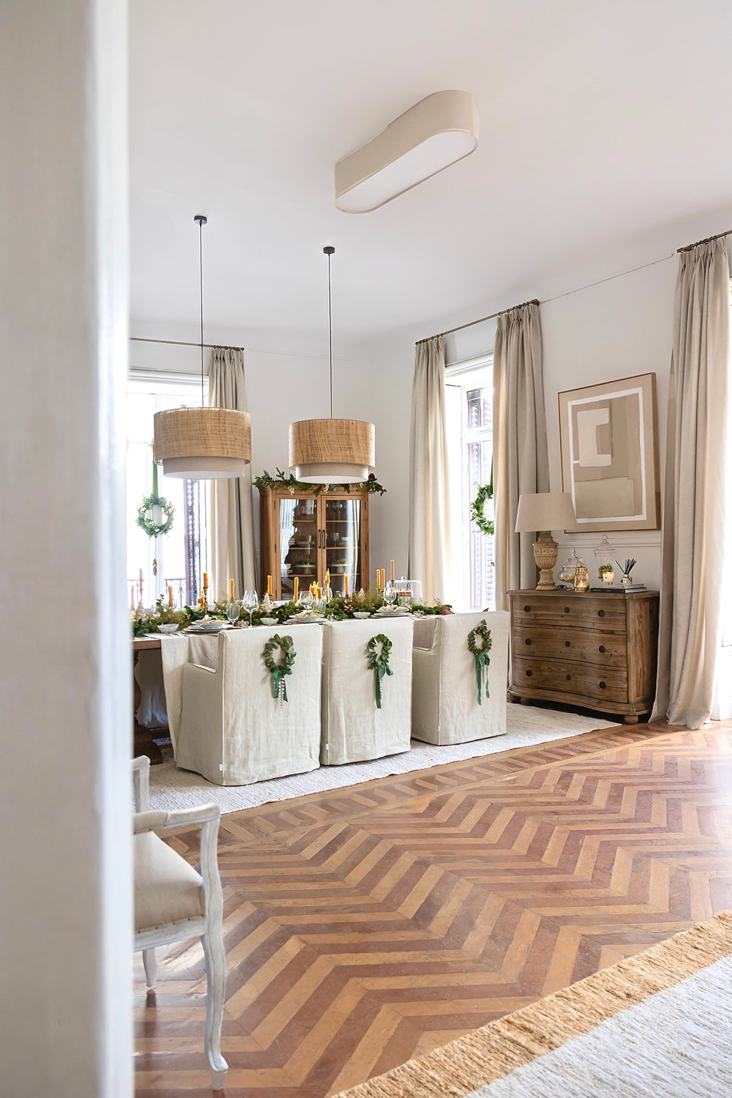 Comedor elegante decorado para Navidad con estilo nórdico, presenta sillas tapizadas en blanco, lámparas colgantes de ratán, piso de madera en espiga, cortinas beige y pequeñas coronas navideñas decorativas