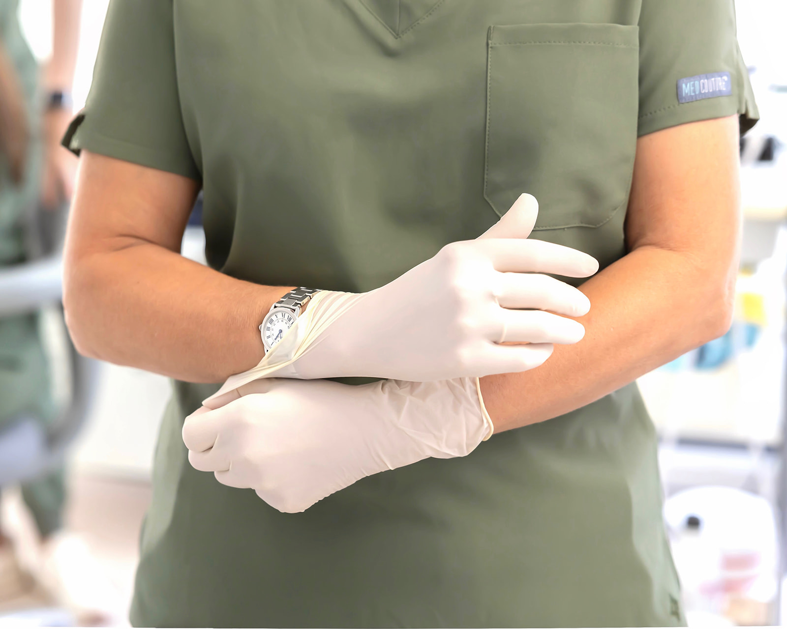 Imagen de un profesional médico vistiendo uniforme verde y colocándose guantes de látex blancos. Se ve un reloj en la muñeca y un bolsillo en el uniforme con una etiqueta.