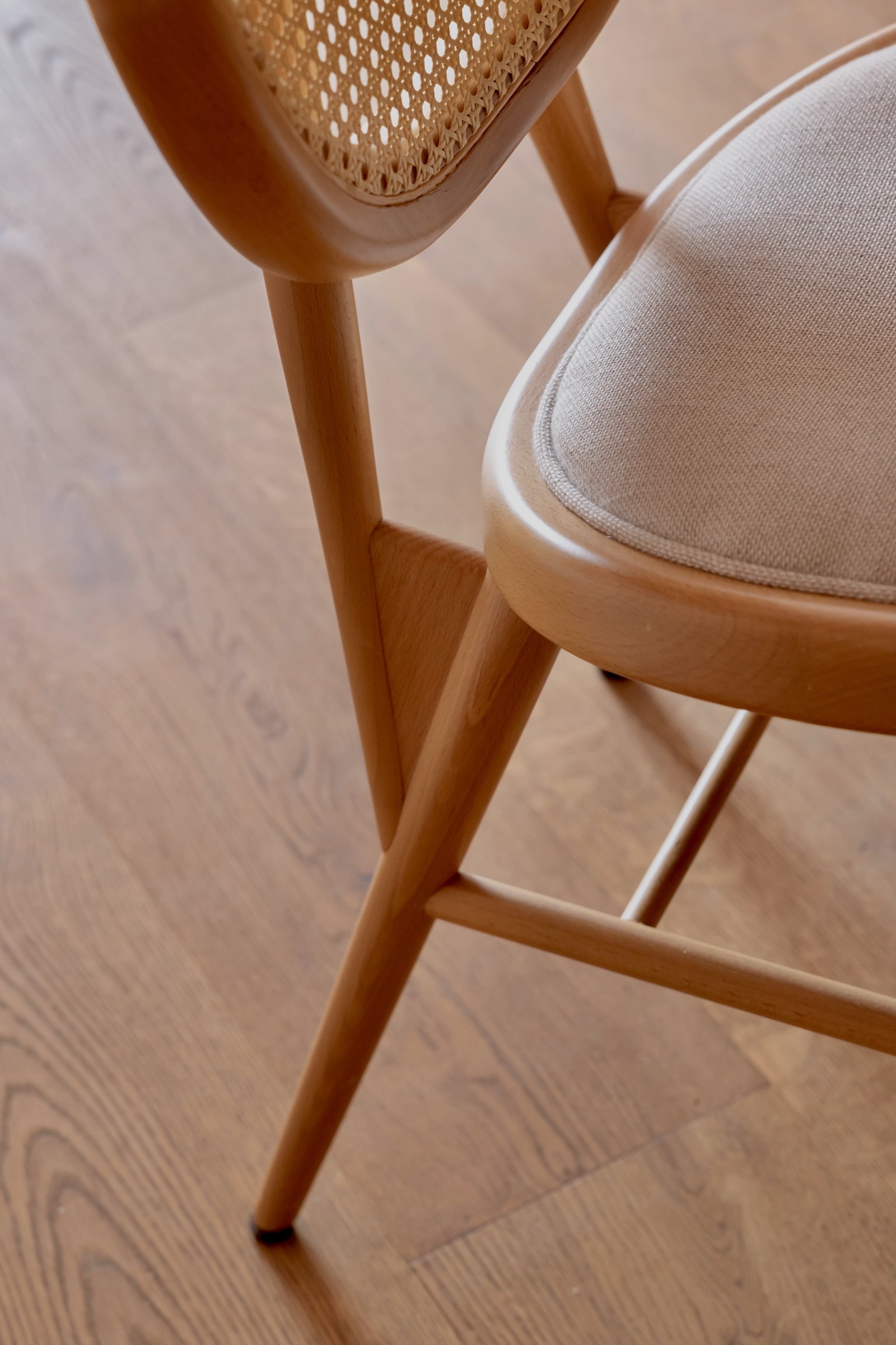 Detalle de silla de madera con asiento beige y respaldo de rejilla, mostrando patas delgadas y estructura elegante sobre piso de madera