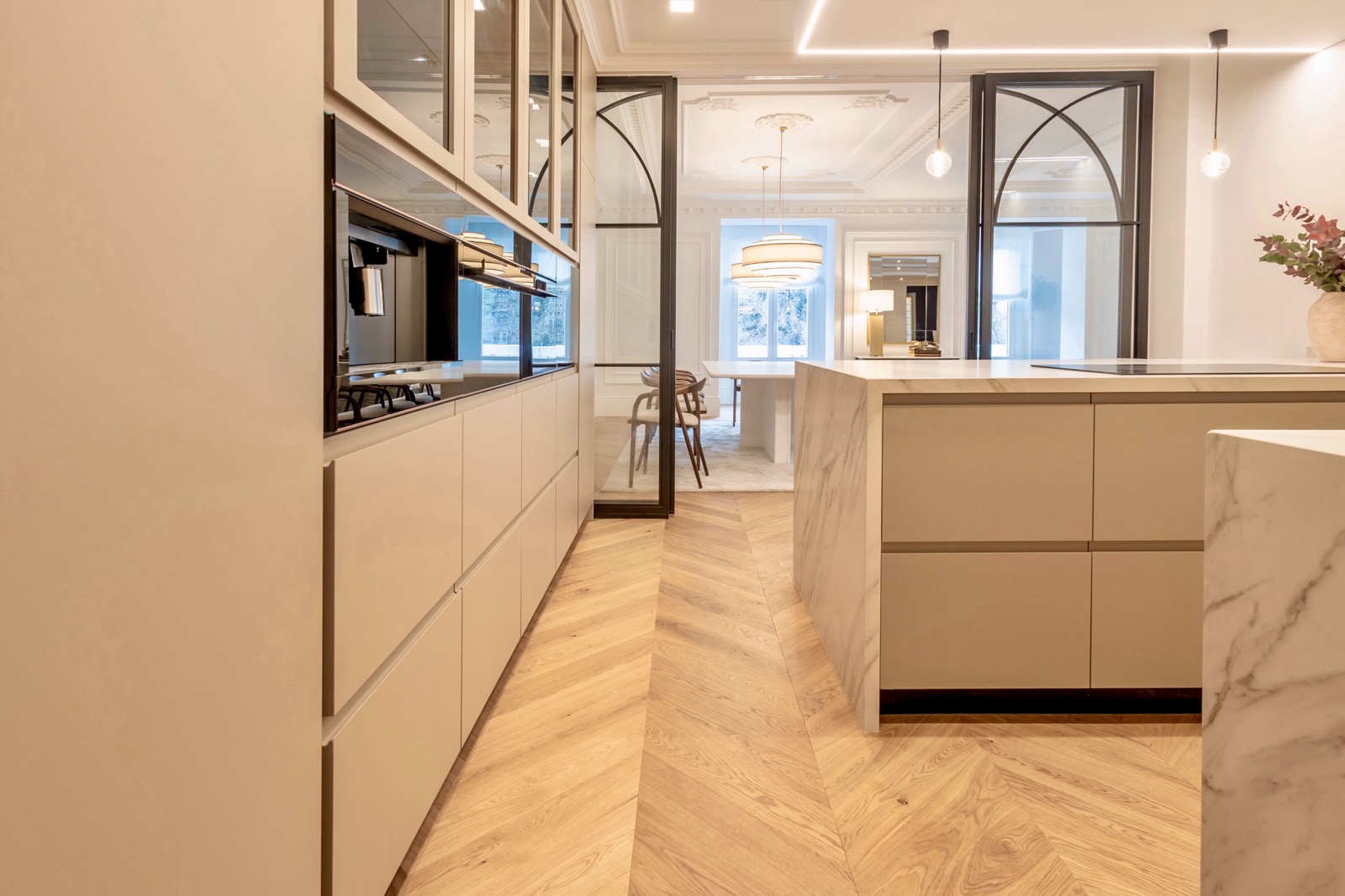 Cocina moderna y elegante con isla de mármol, gabinetes beige, piso de madera en espiga, y puertas de vidrio que separan el comedor. Iluminación empotrada y colgante crea un ambiente sofisticado.