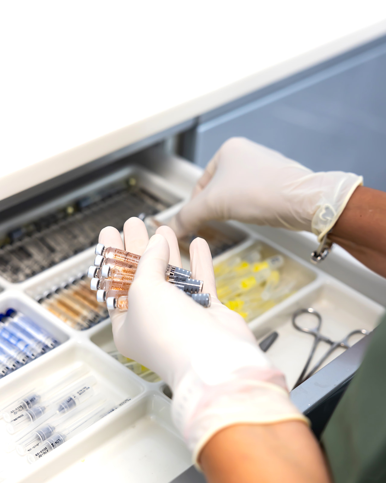 Manos con guantes médicos organizando viales y suministros en un cajón de almacenamiento médico. Se ven diversos instrumentos y materiales médicos en compartimentos separados.