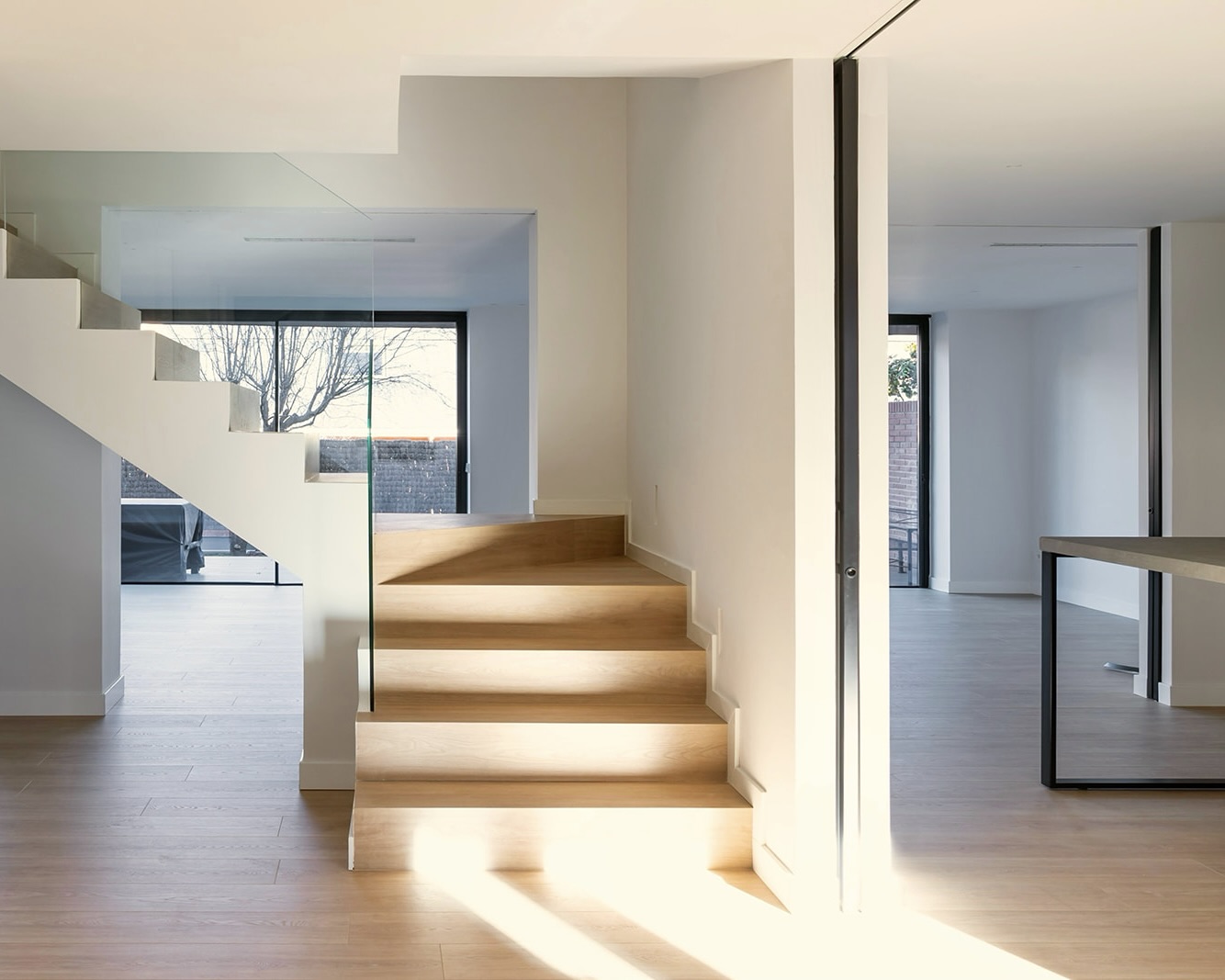 Interior moderno con escaleras de madera iluminadas, paredes blancas, ventanas grandes y piso de madera. Diseño minimalista con elementos de vidrio y metal, creando un espacio luminoso y abierto.