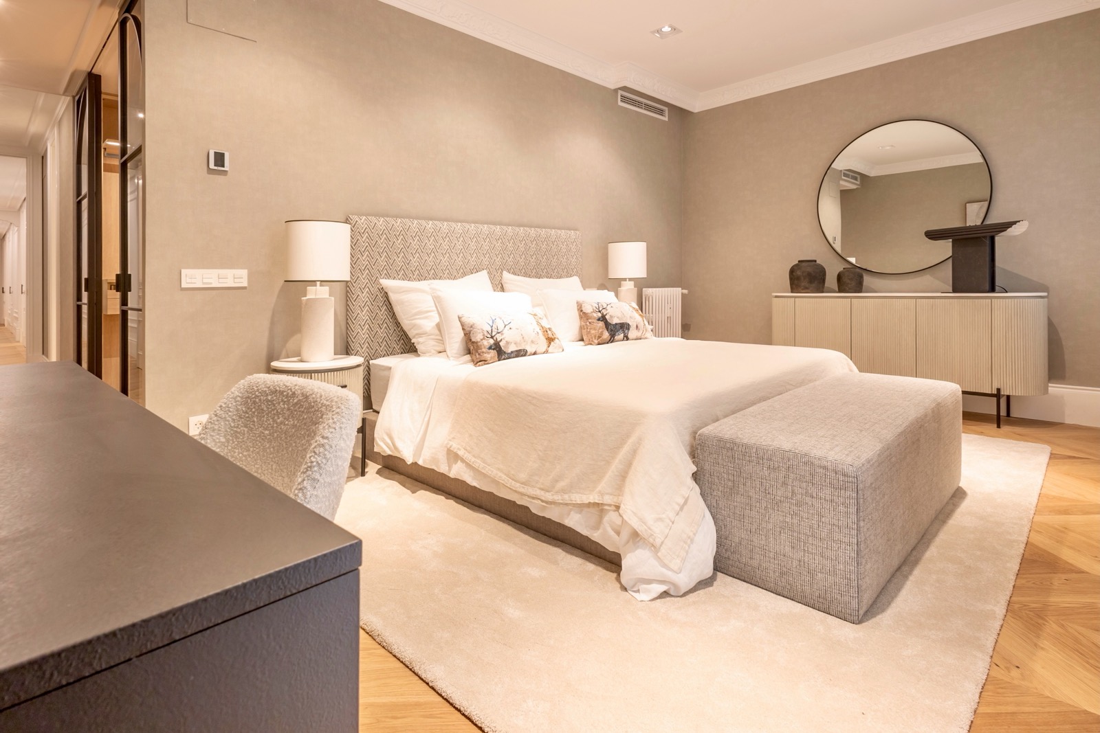 Dormitorio elegante con cama grande, paredes beige, espejo redondo, cómoda y decoración en tonos neutros. El diseño es moderno y minimalista, con iluminación suave y acogedora.