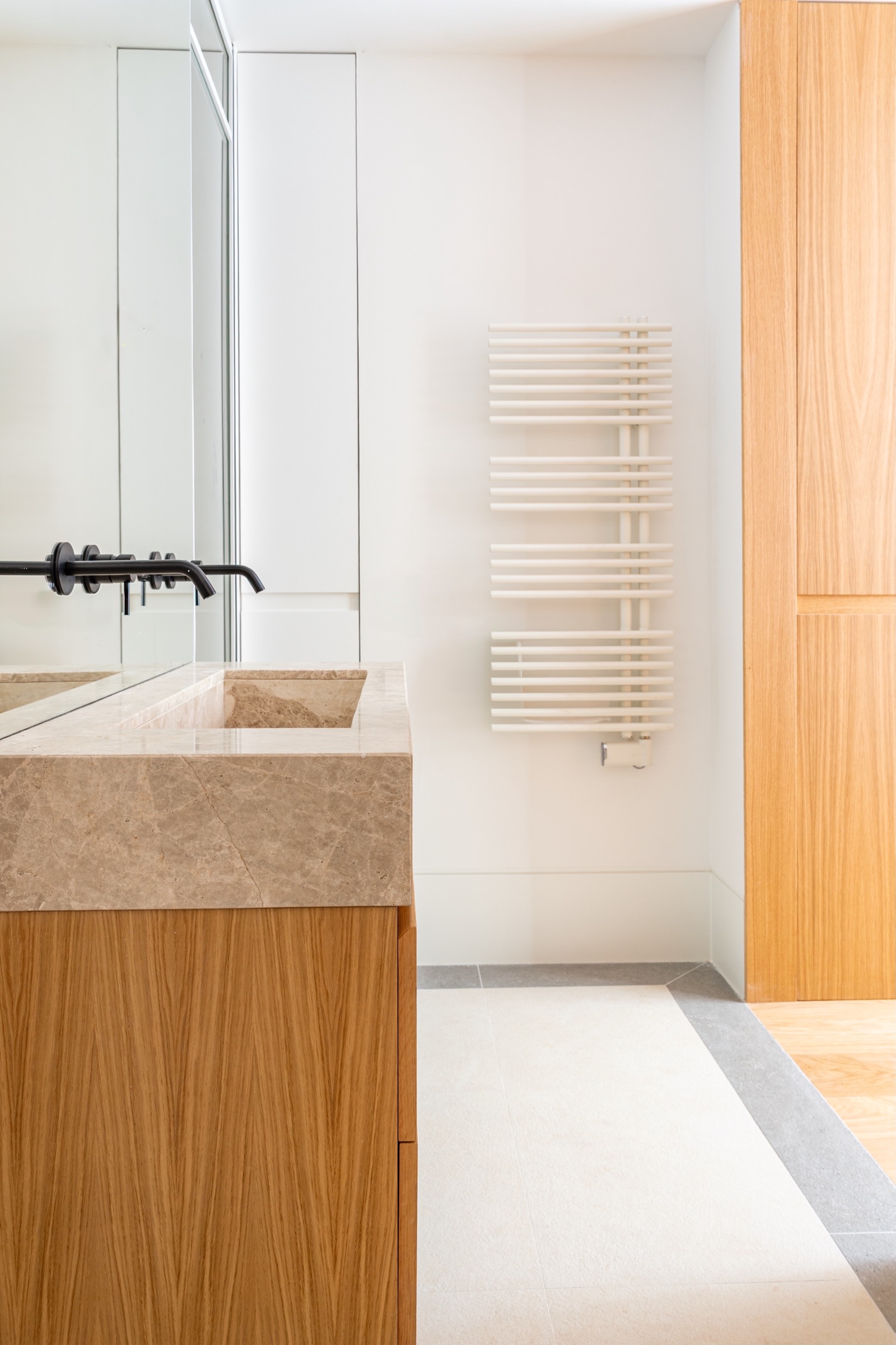 Detalle de lavamanos de mármol beige con mueble de madera, espejo y toallero blanco en un baño minimalista